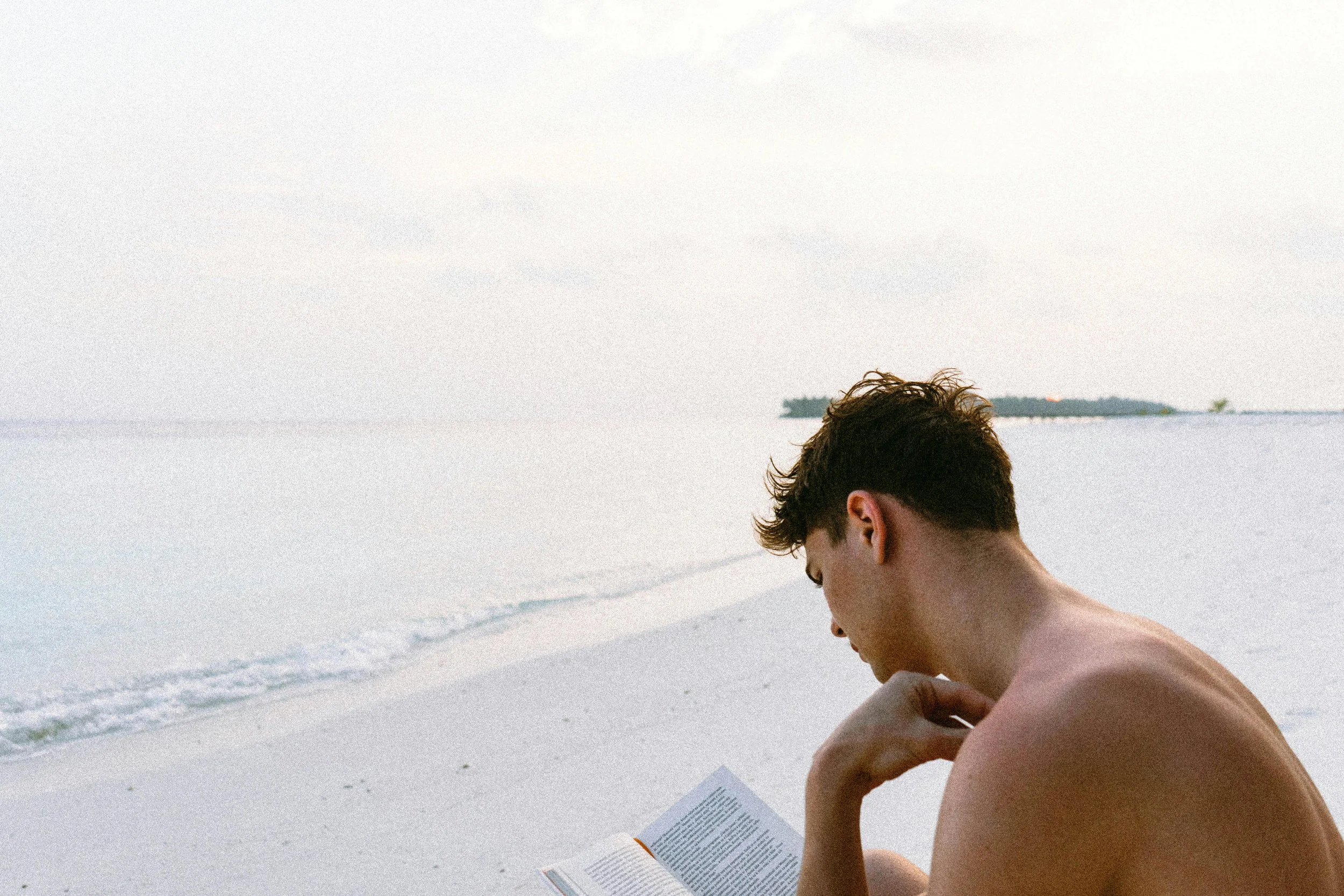 A young man reading a book on the beach with a calm ocean and distant island in the background.