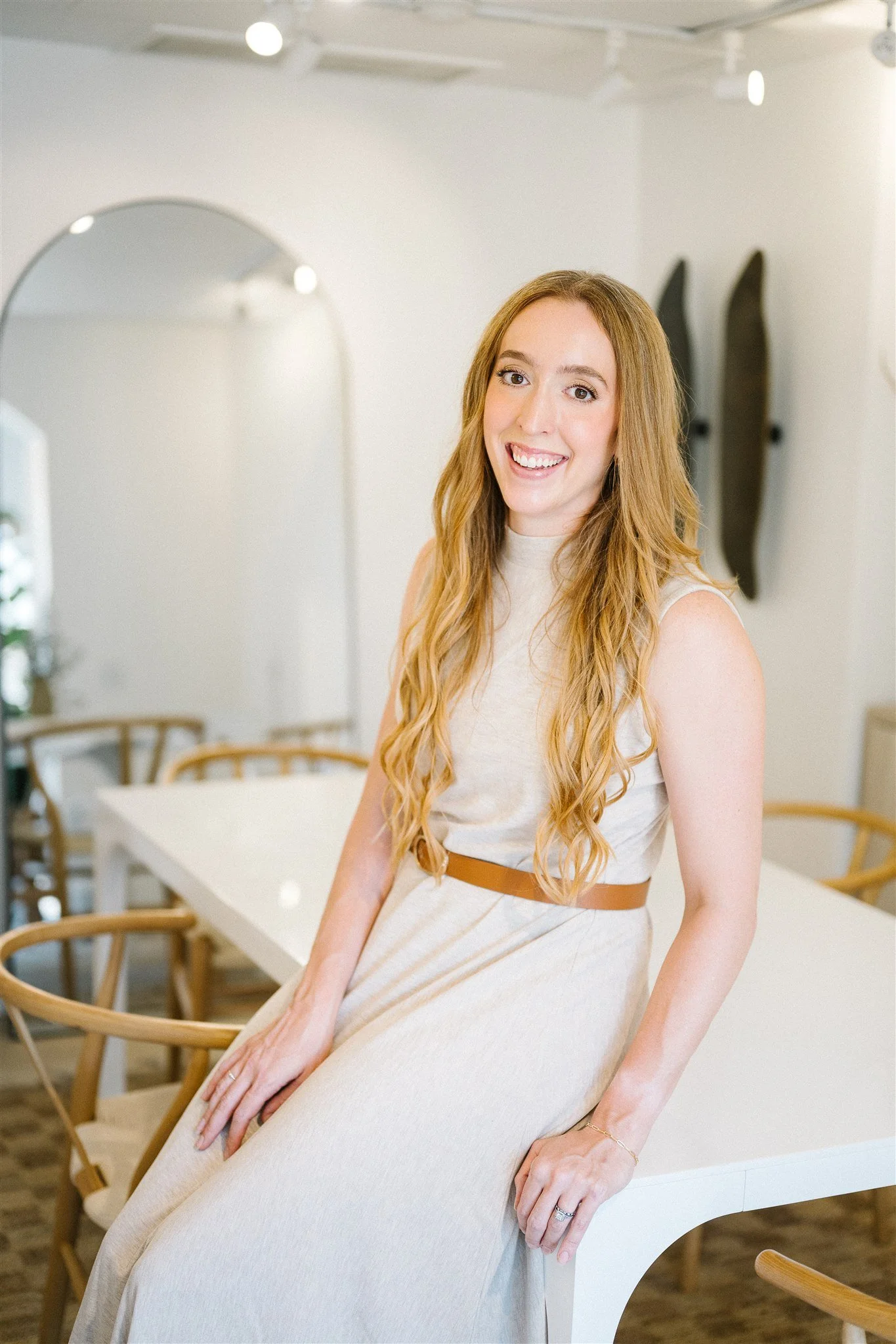 A woman with long, wavy blonde hair sitting on a white dining table in a modern, bright room with wooden chairs and skateboards mounted on the wall in the background.