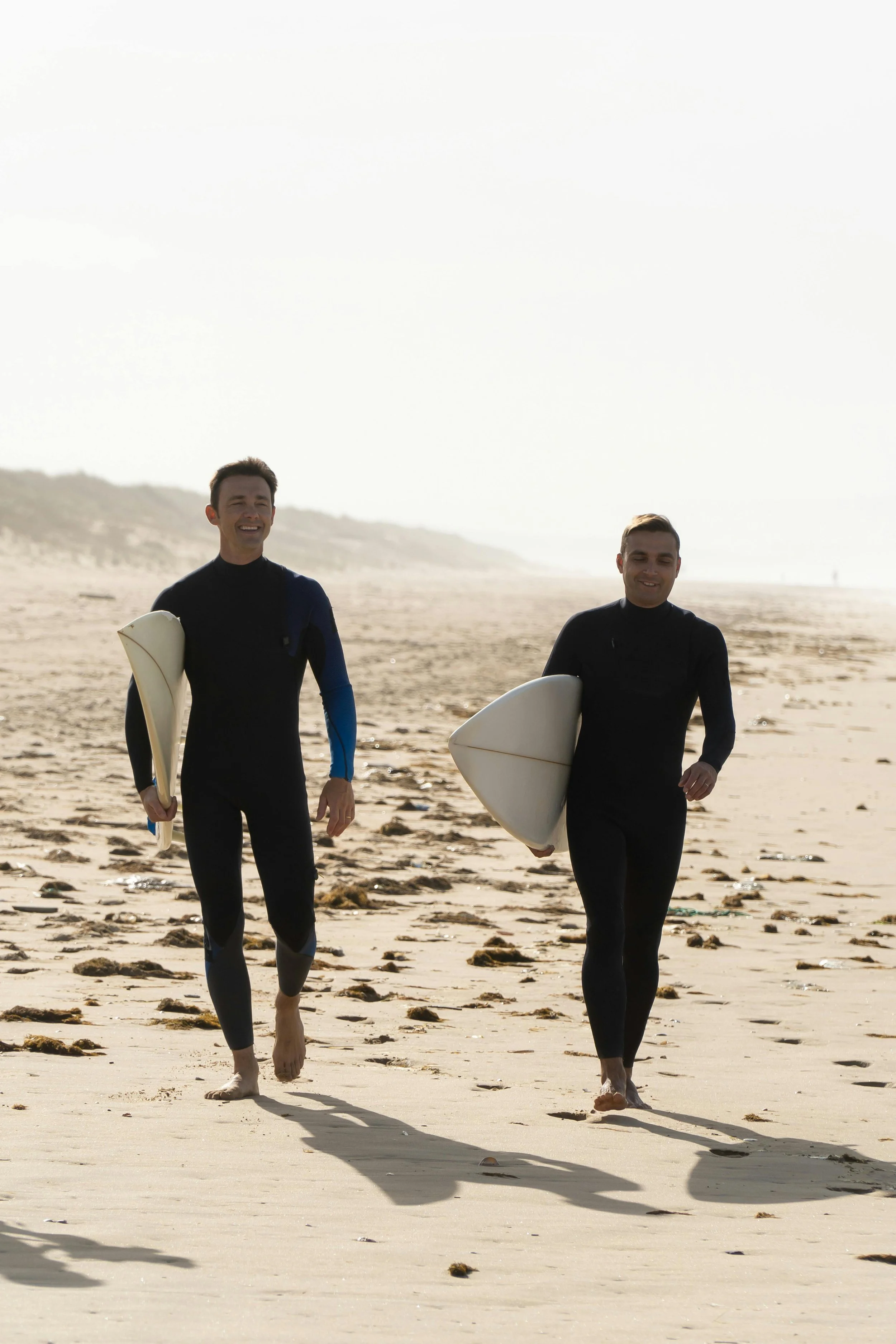 Two men in wetsuits walking on a beach carrying surfboards, smiling and enjoying the day.