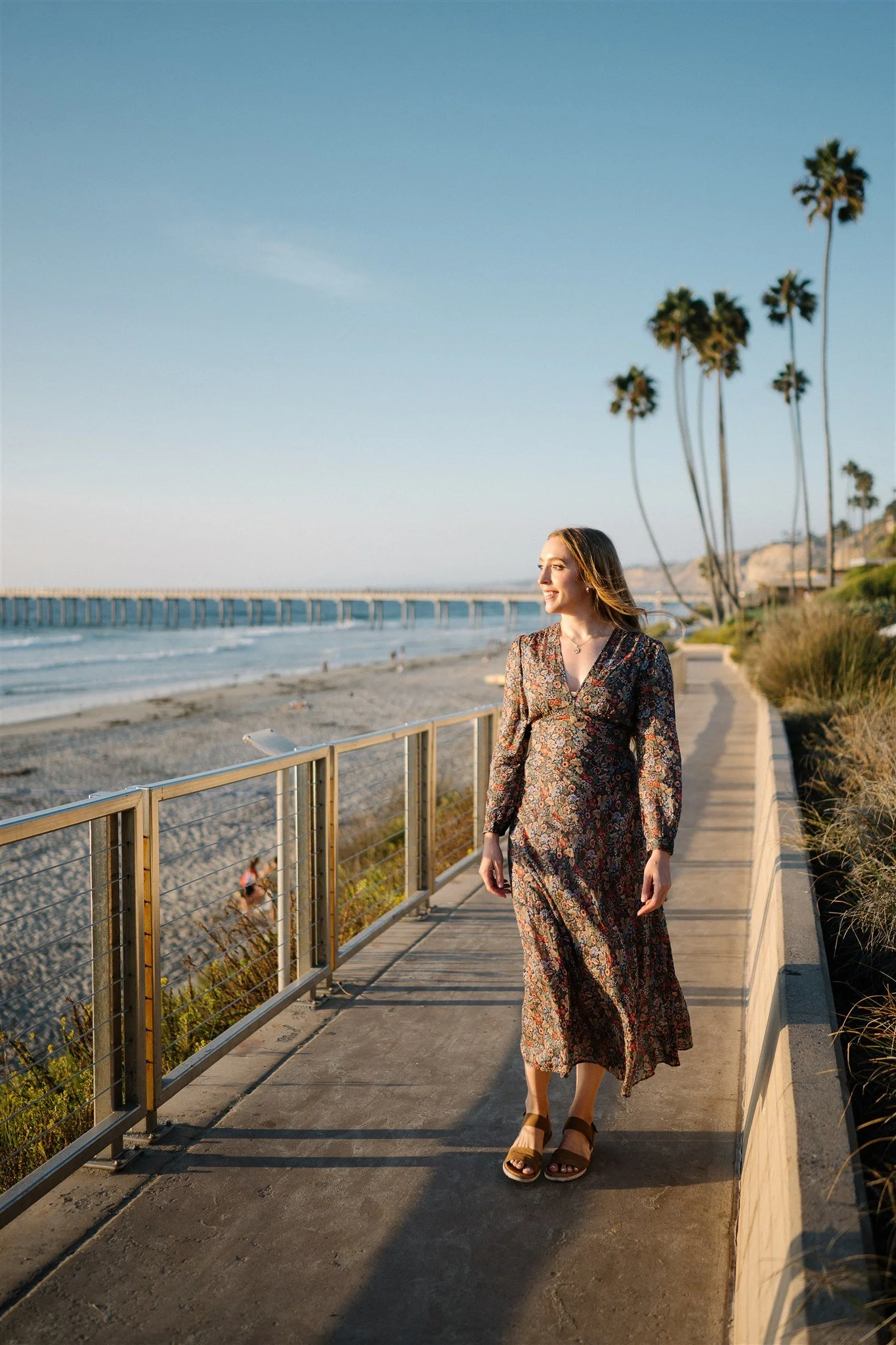A woman in a floral dress walking along a seaside path with palm trees, ocean, beach, and a pier in the background during sunset.