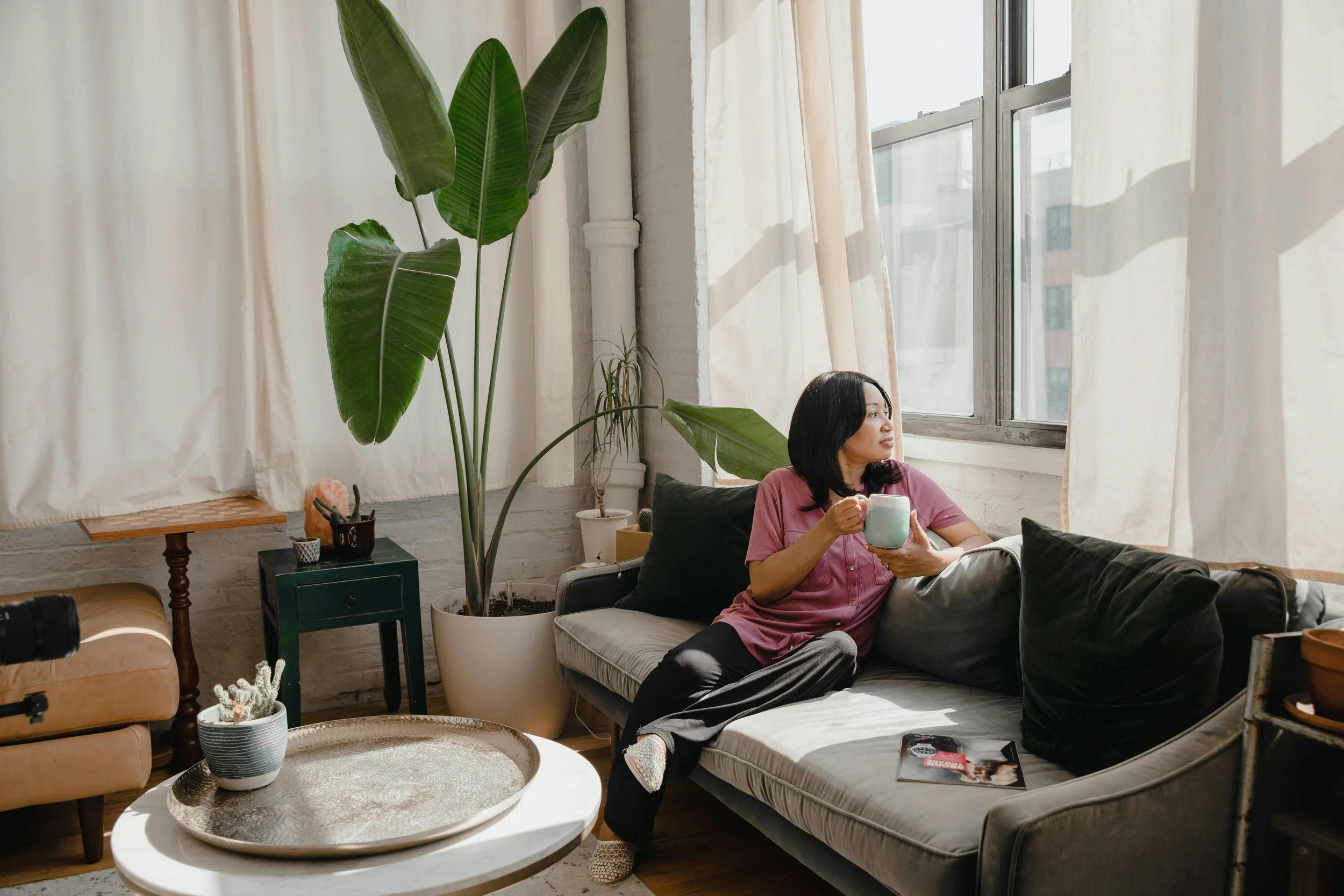 Woman sitting on a beige sofa near a large window, holding a light green mug, with sunlight streaming in. The living room has potted plants, a round coffee table with a decorative tray, and a magazine on the sofa.