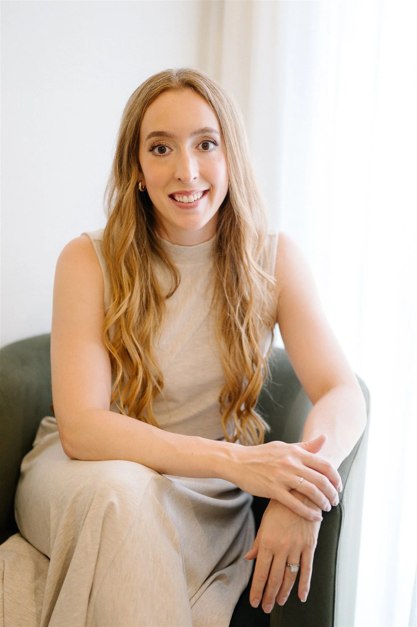 A woman with long, wavy red hair sitting on a green armchair near a window with sheer curtains, smiling at the camera.