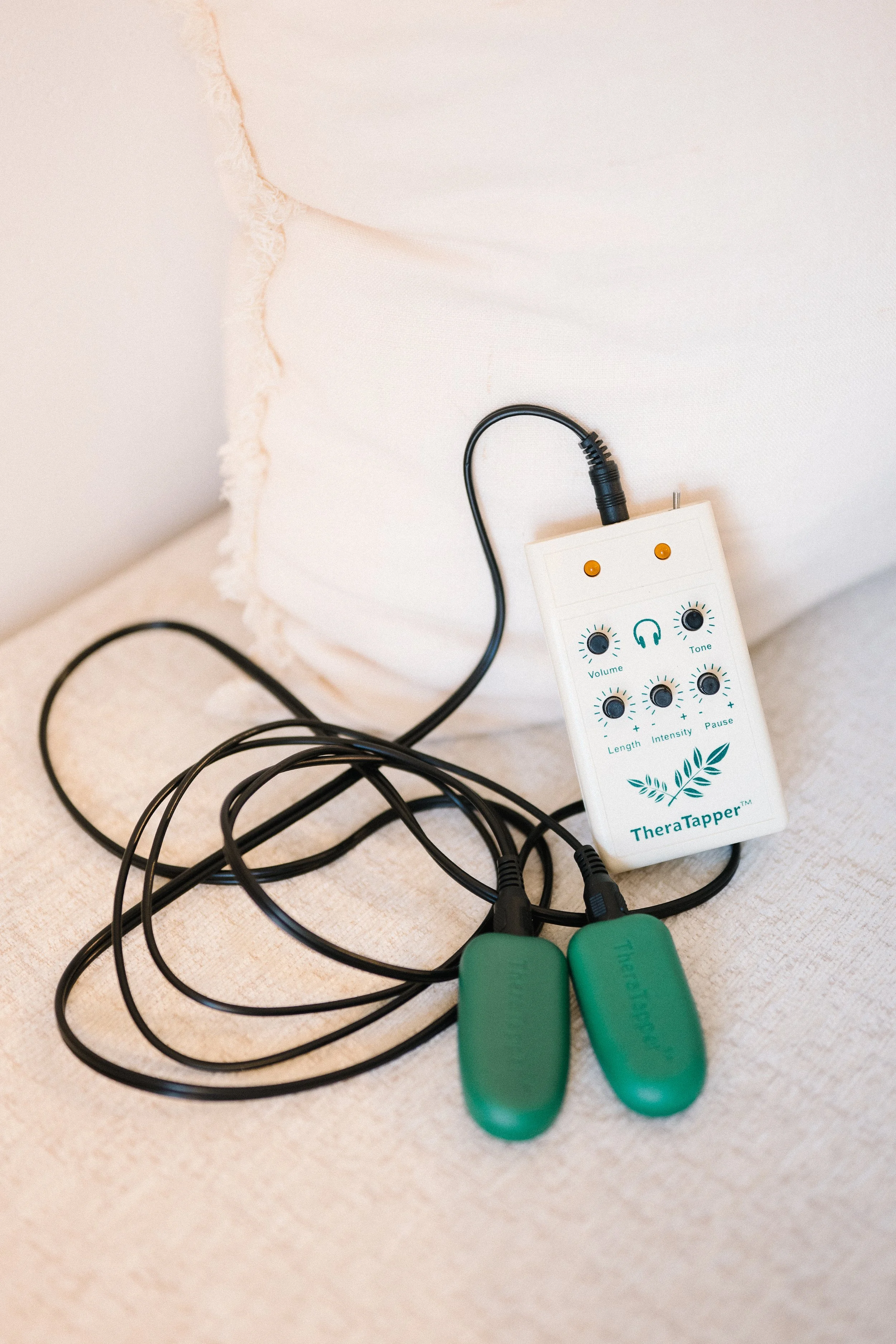 A TheraTapper device connected to two green therapy paddles, sitting on a beige fabric surface near a white pillow.