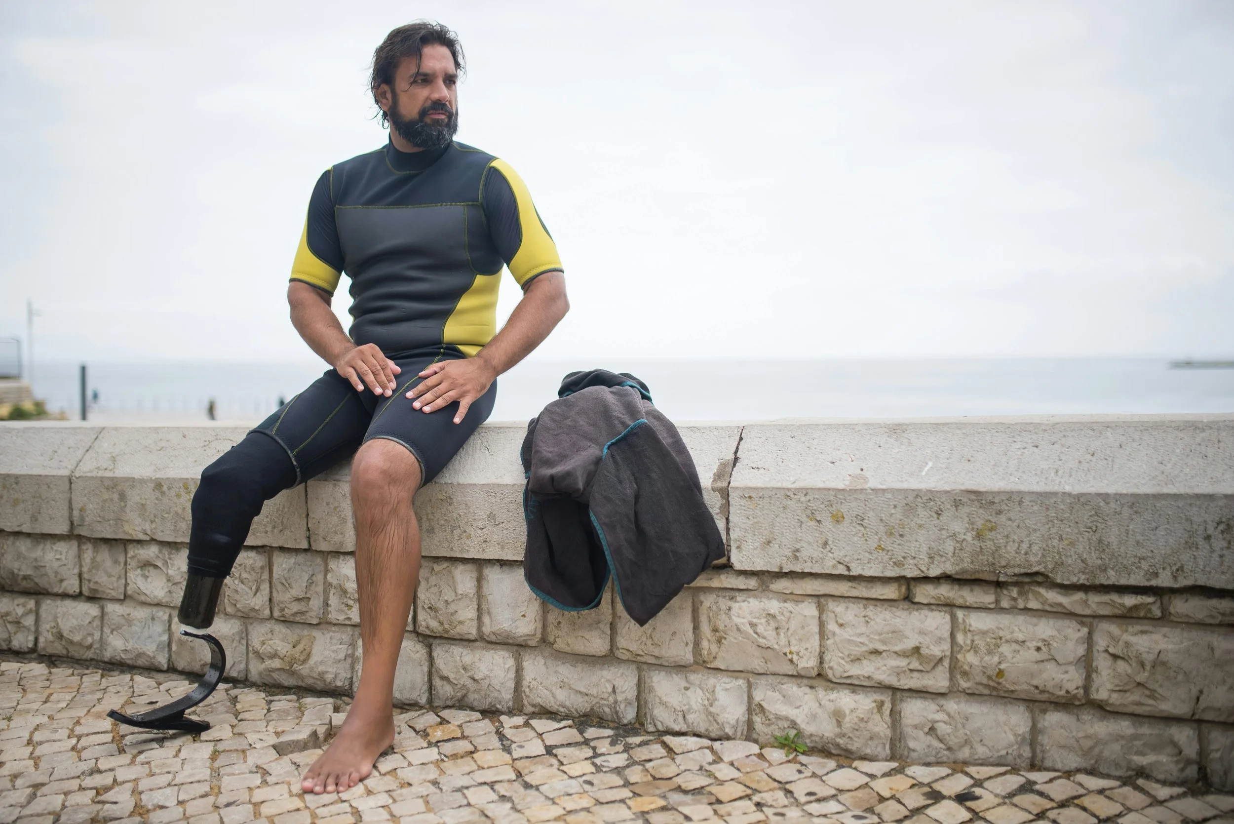 A man with a prosthetic leg wearing a wetsuit, sitting on a stone barrier by the sea with a backpack next to him, looking to his left on a cloudy day.
