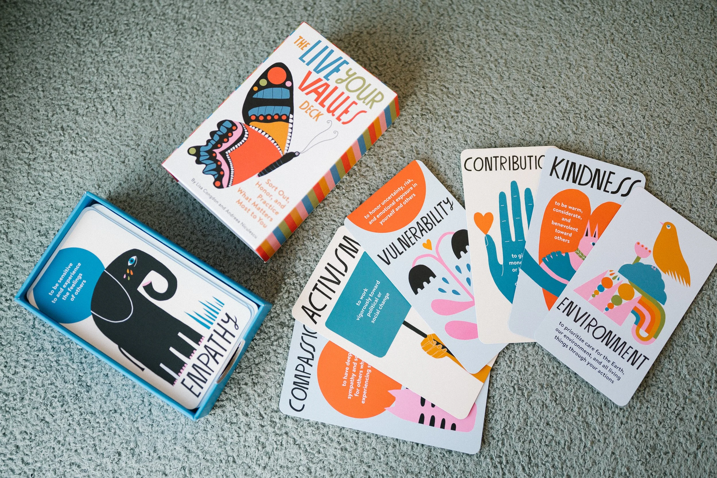 Cards and a box for a game called 'The Live Your Values Deck' with colorful illustrations and words like empathy, compassion, vulnerability, contribution, kindness, and environment, placed on a gray carpet.