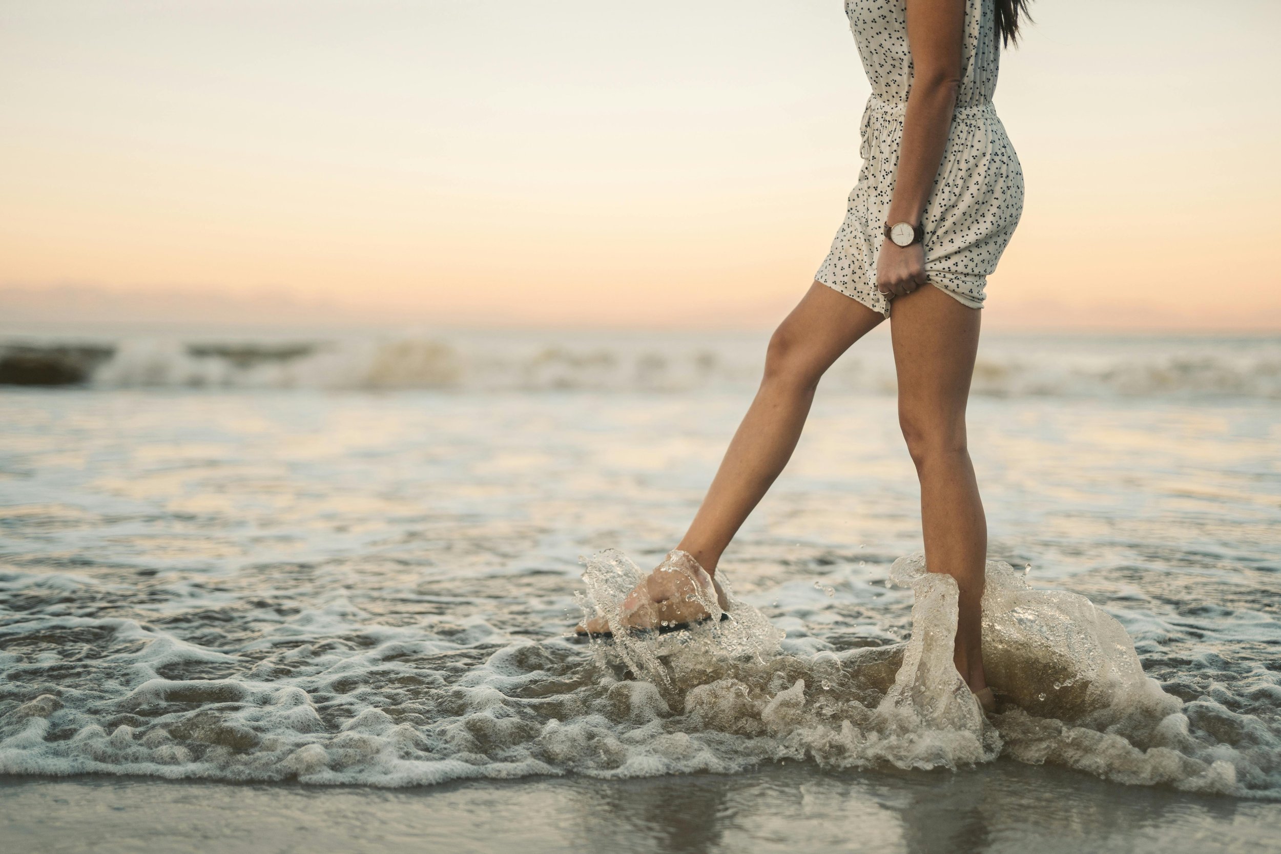 A woman in patterned summer romper splashing water at the beach during sunset.