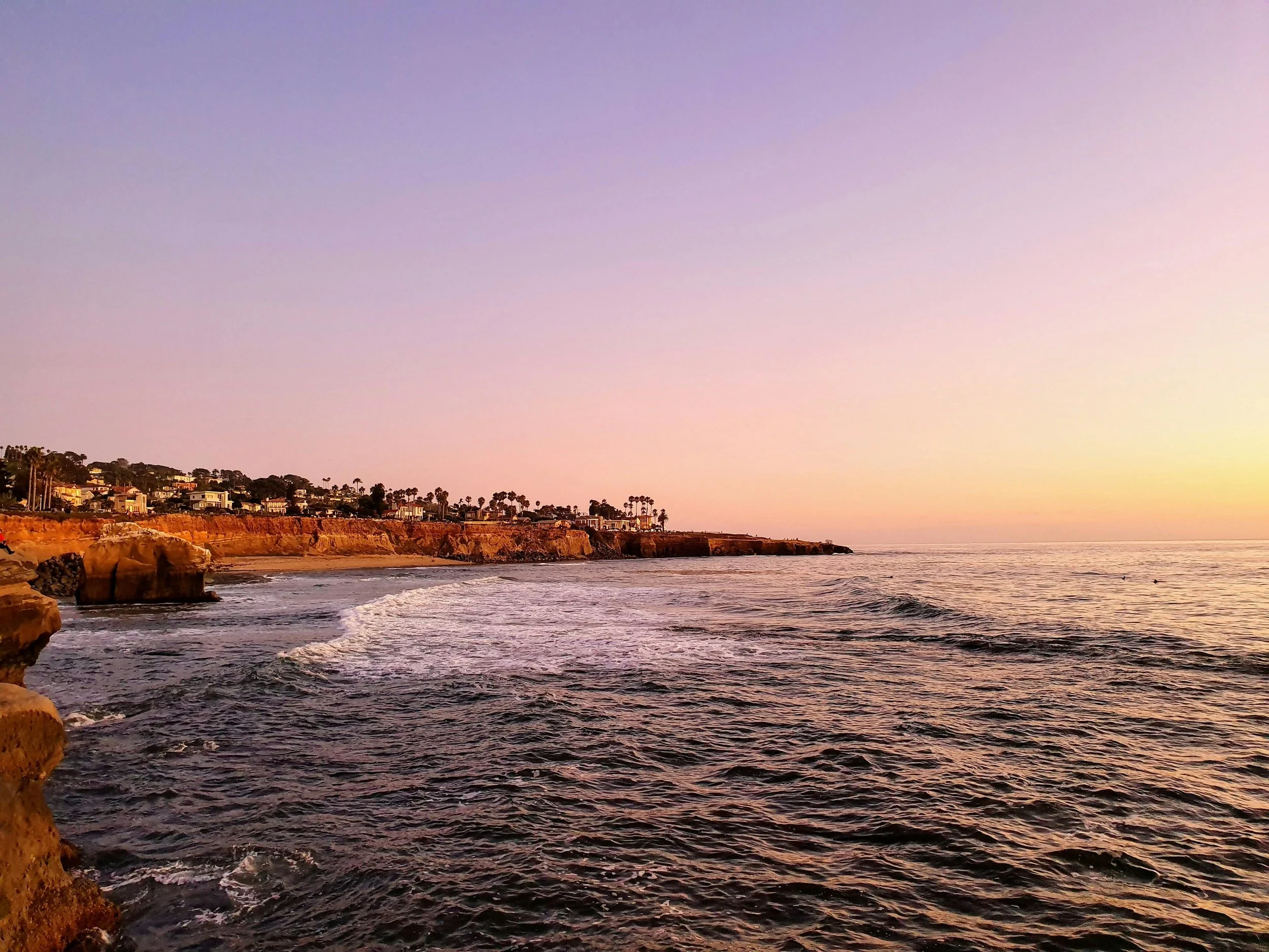 Sunset over the ocean with rocky cliffs and a coastal town in the background.