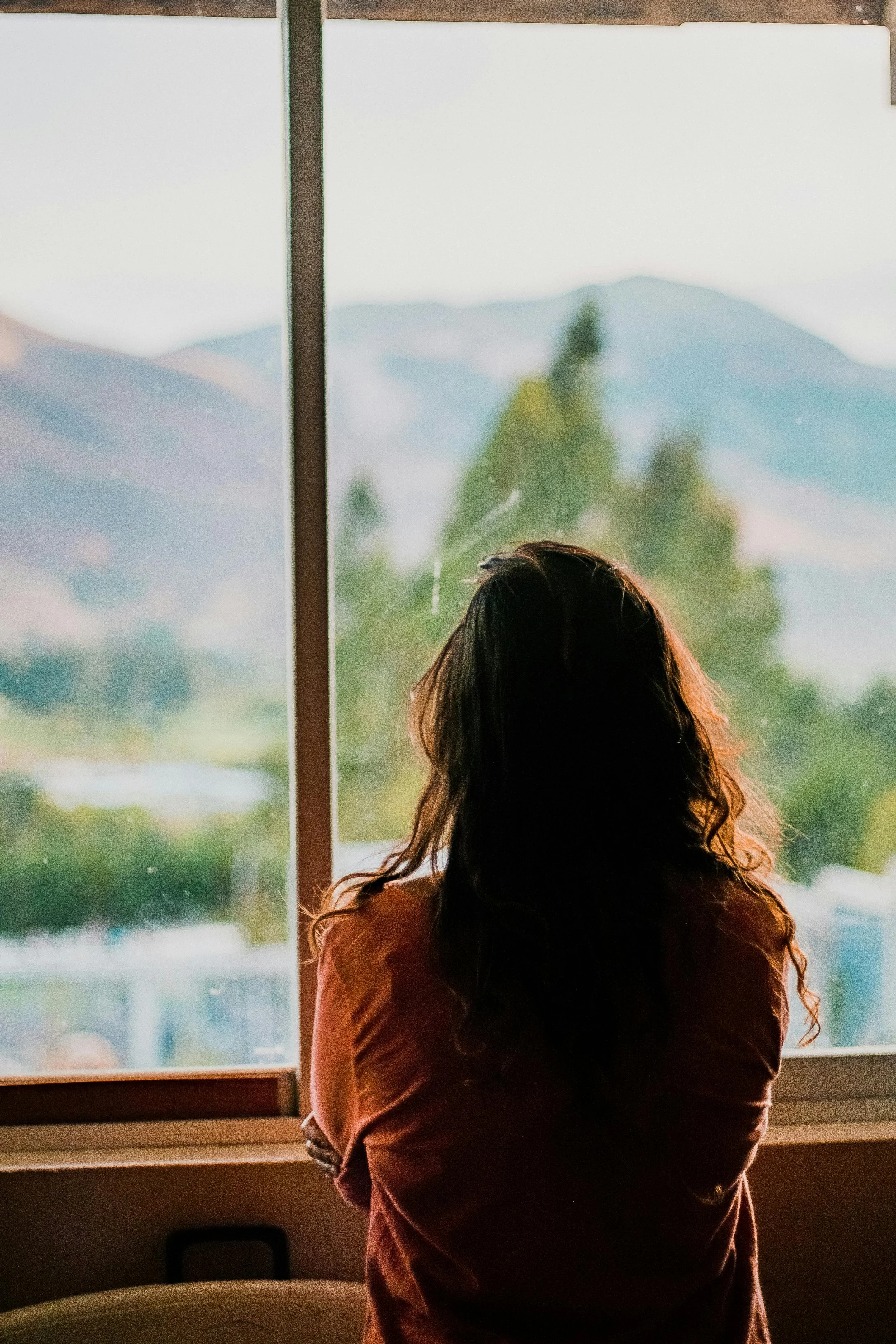 A woman with long, wavy hair standing indoors, looking out a window at a mountainous landscape with trees and a cloudy sky.