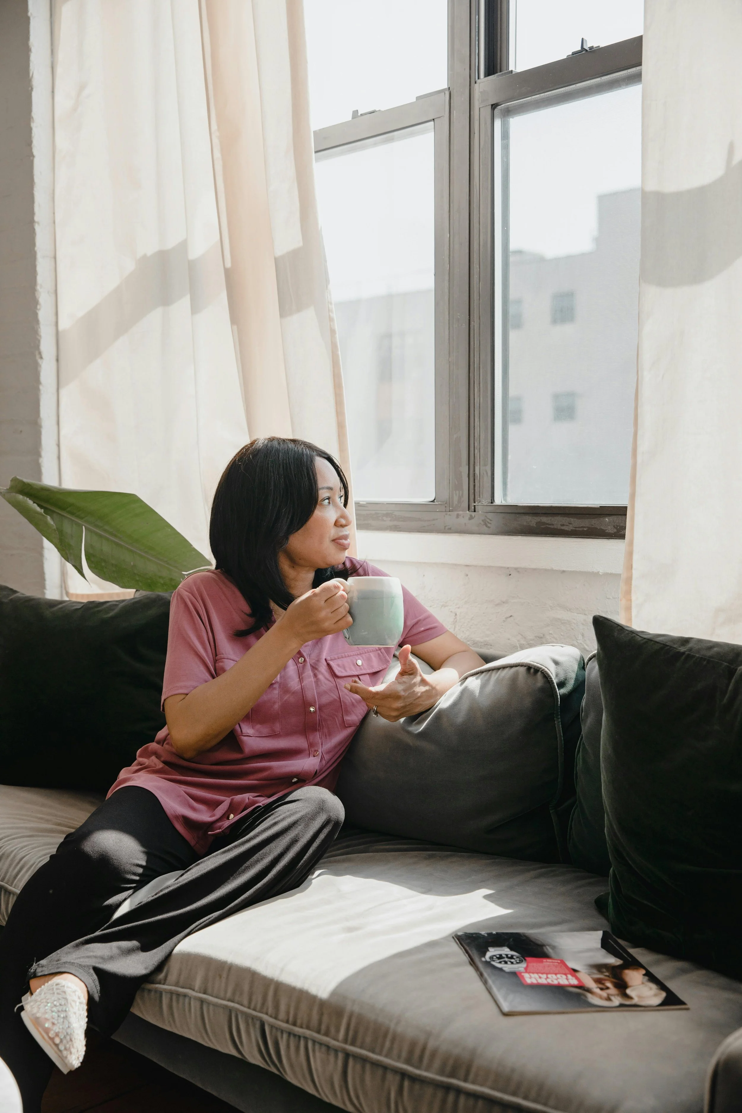 A woman sitting on a sofa near a window, holding a mug, with magazines or books on the sofa, and a large green leaf plant behind her.