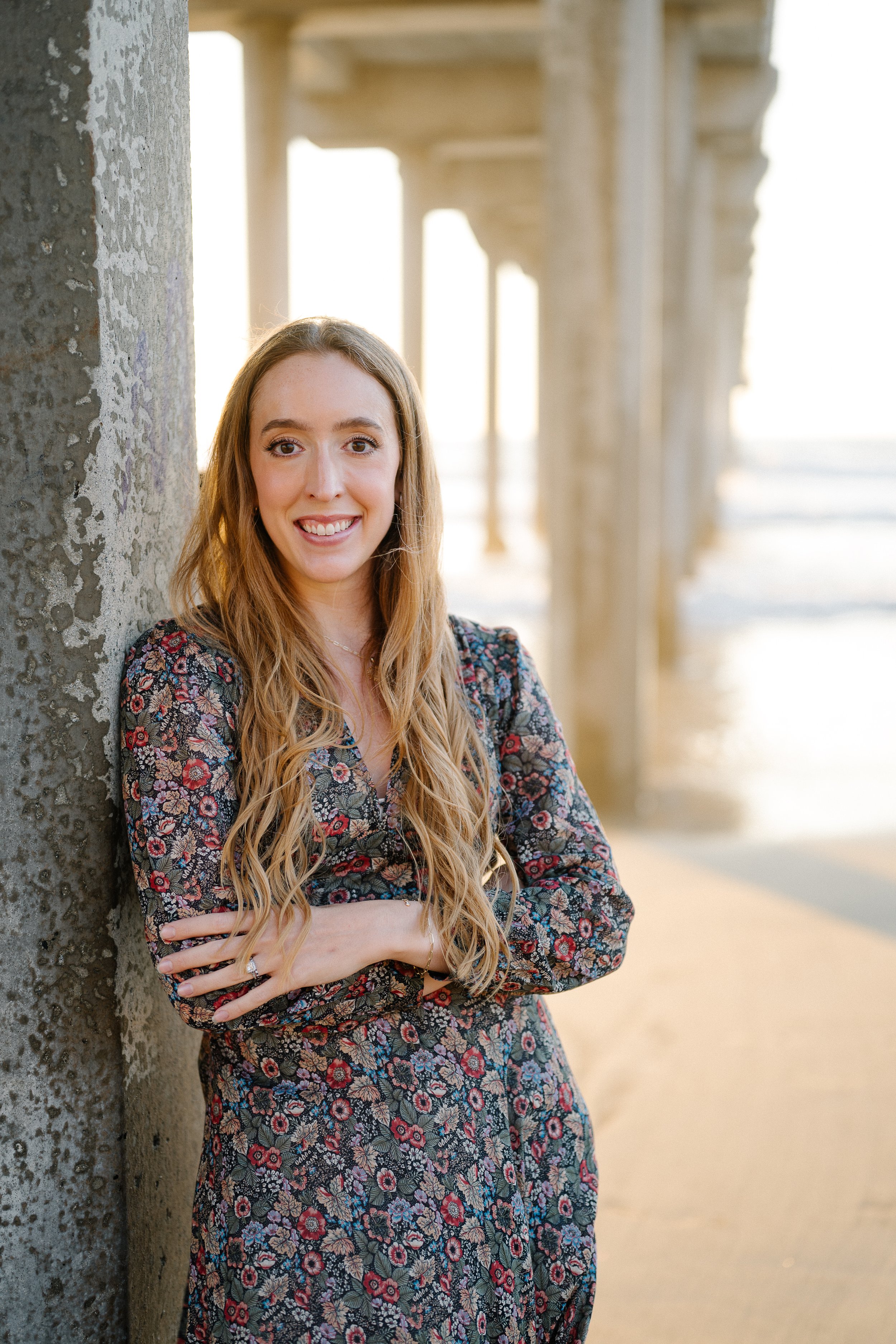 A young woman with long, wavy blonde hair standing on a beach under a pier. She is leaning against a concrete pillar, smiling, wearing a dark floral dress with long sleeves.