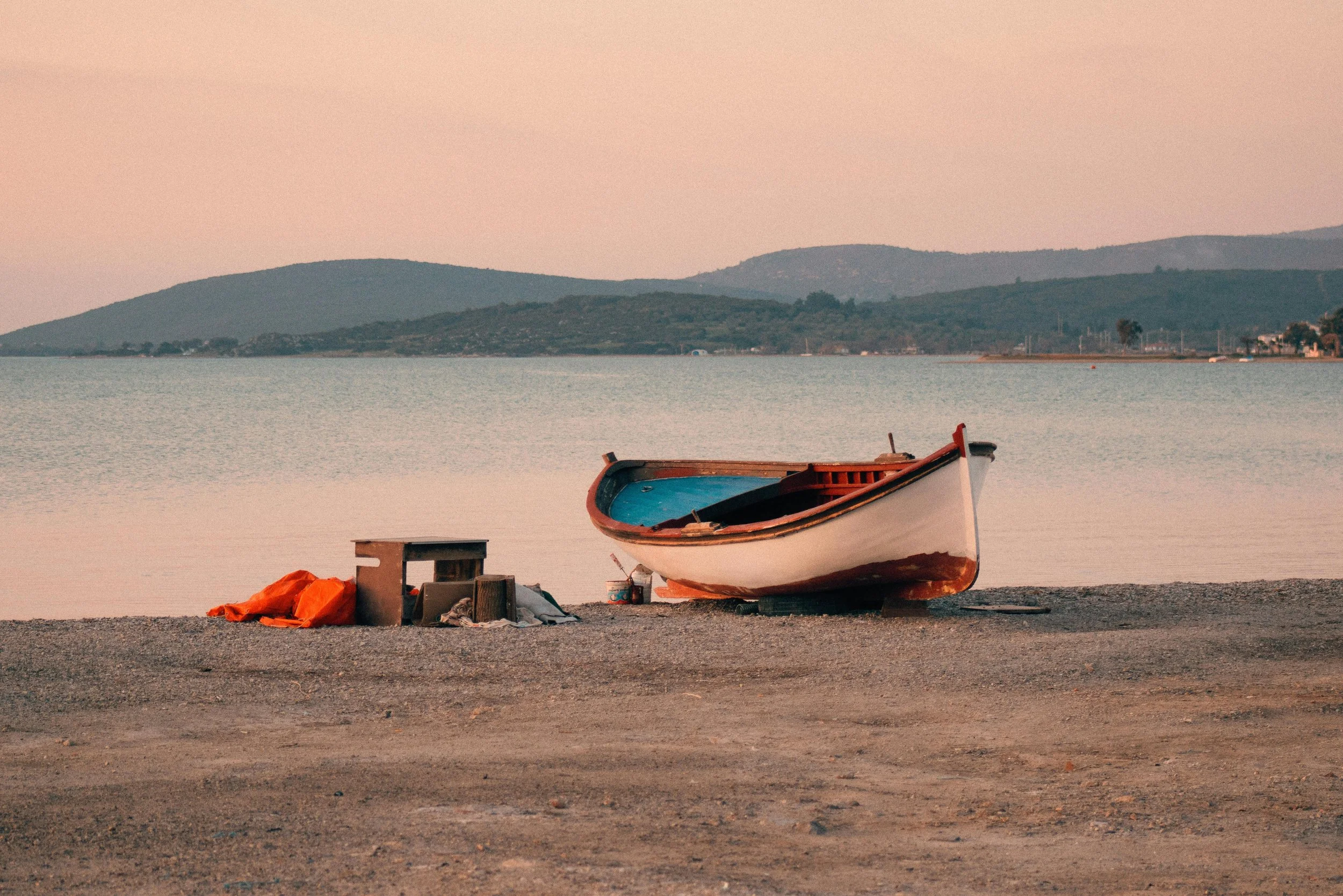A small boat resting on the shore of a calm lake or ocean, with mountains in the background and a pastel pink sky, suggesting dawn or dusk.