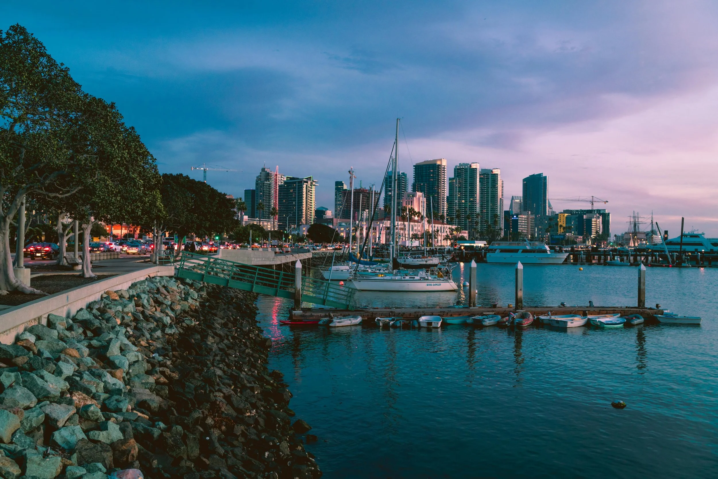 Boats docked at a marina with a city skyline and high-rise buildings in the background during sunset