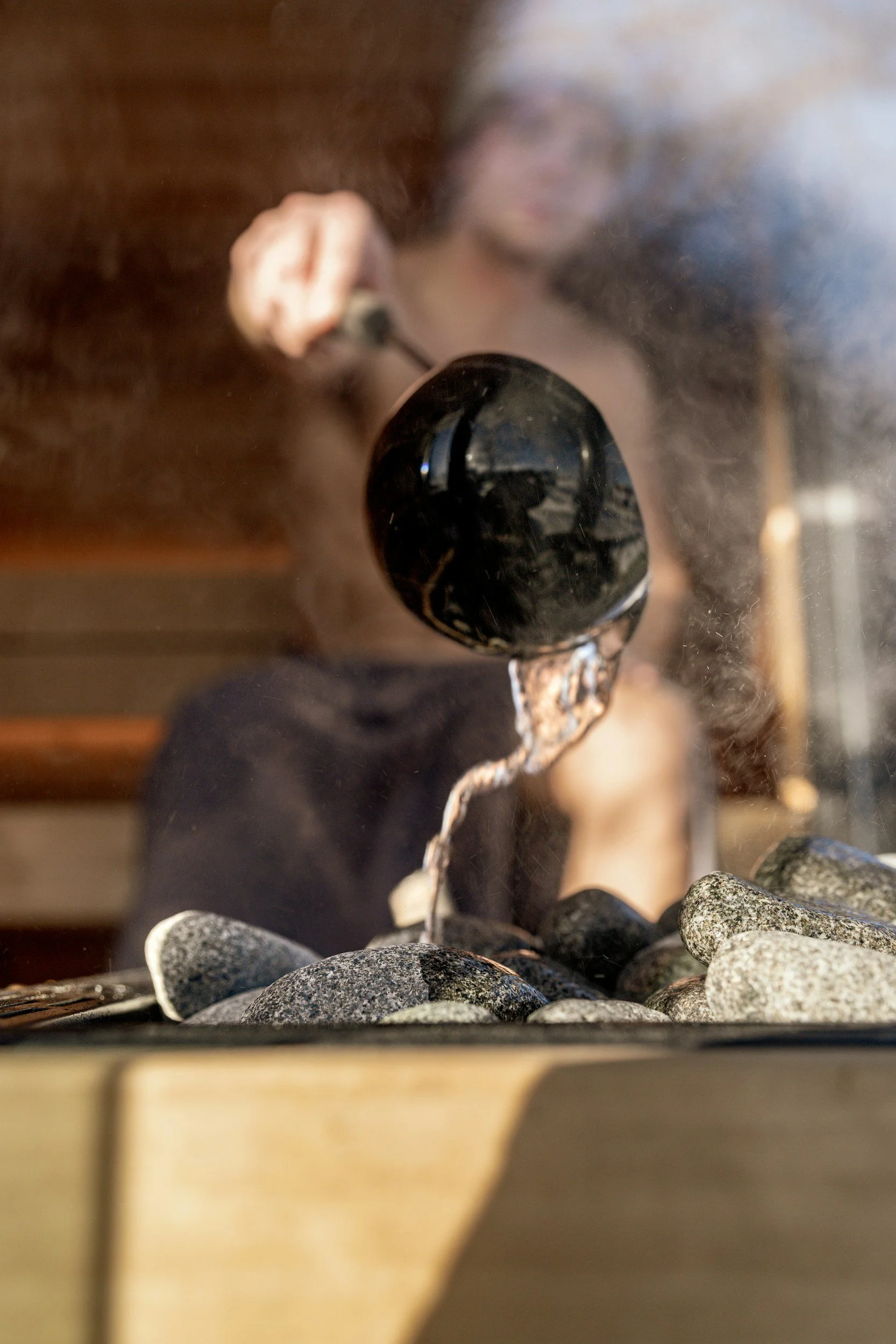 A woman is pouring hot water from a black kettle onto rocks on a table.
