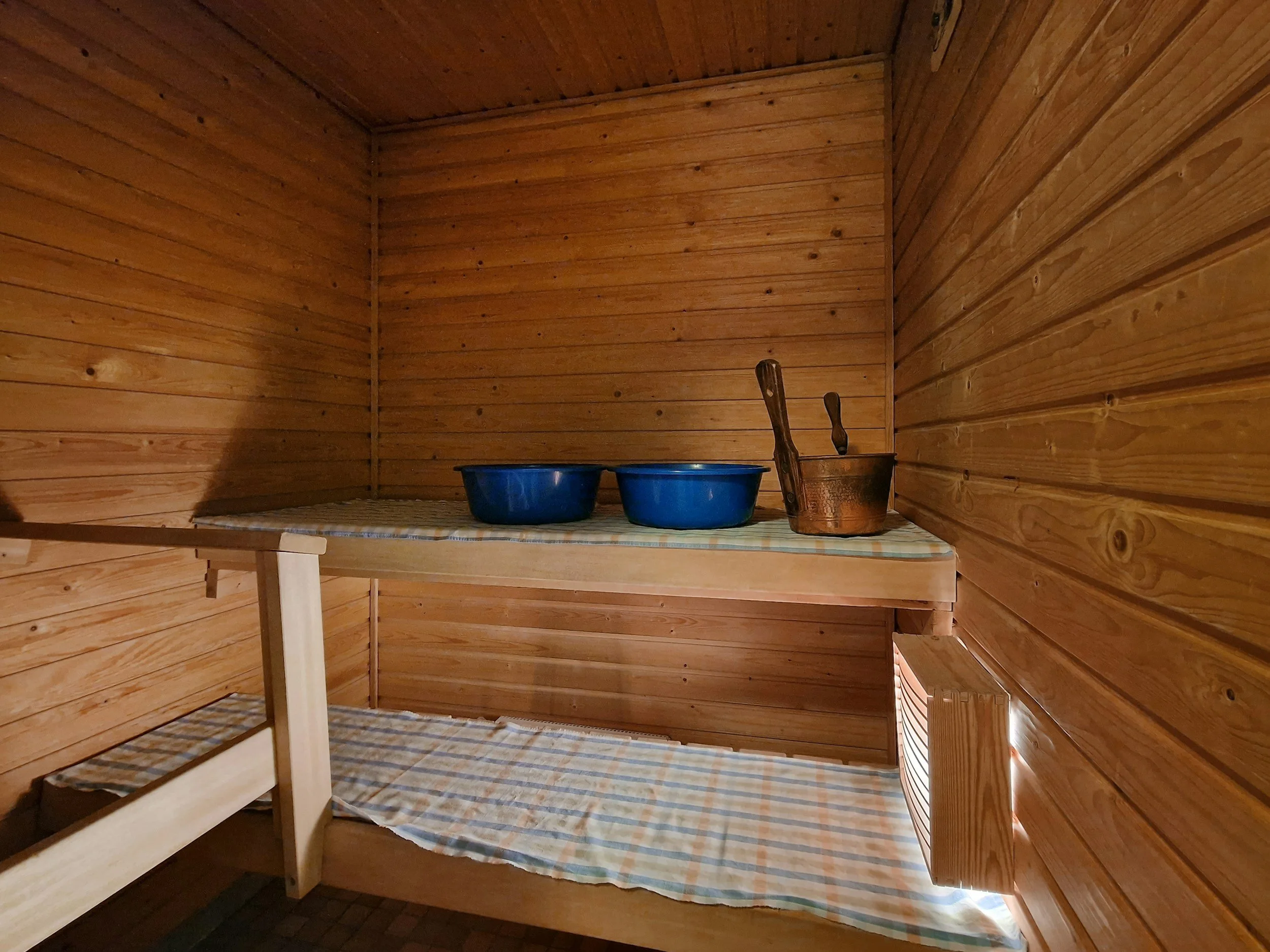 Empty wooden sauna room with two blue buckets and a copper bucket with a ladle on a striped cloth-covered shelf.