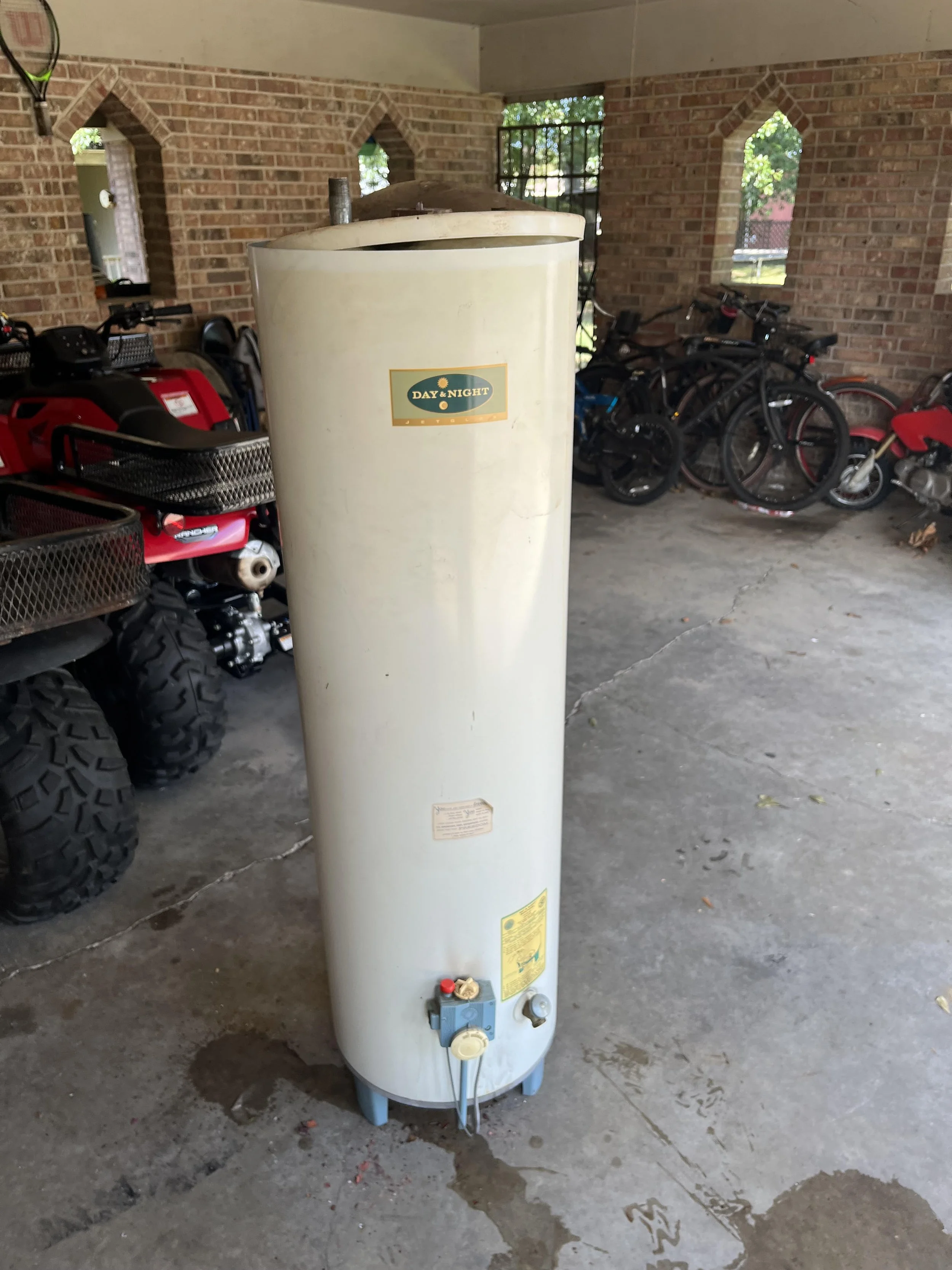 A tall, cylindrical water heater with a label that reads "Day & Night" in a garage with brick walls, and parked bikes and ATV.