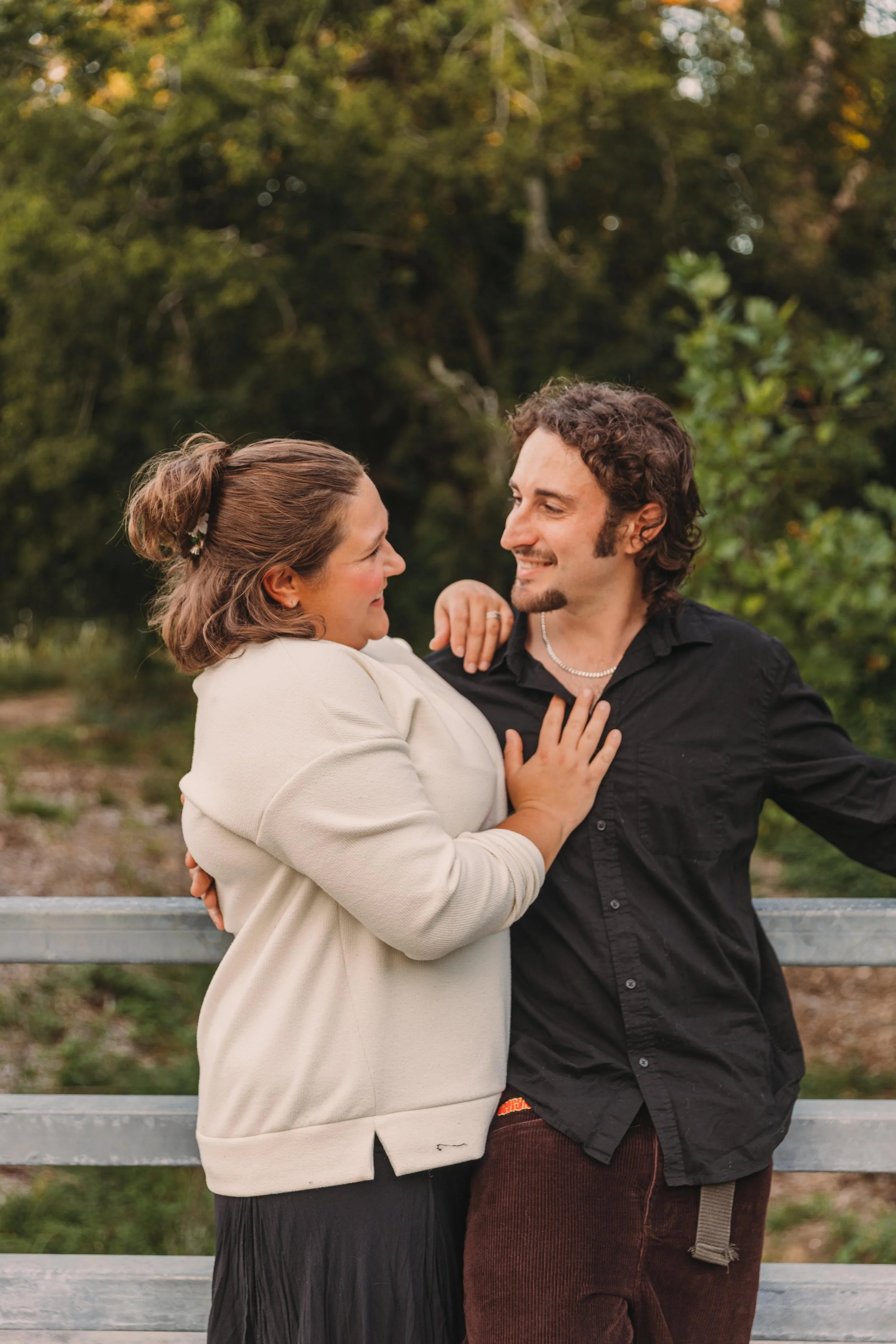 A couple standing close together outdoors, smiling at each other, with the woman touching the man's chest. They are near a metal railing with trees in the background.