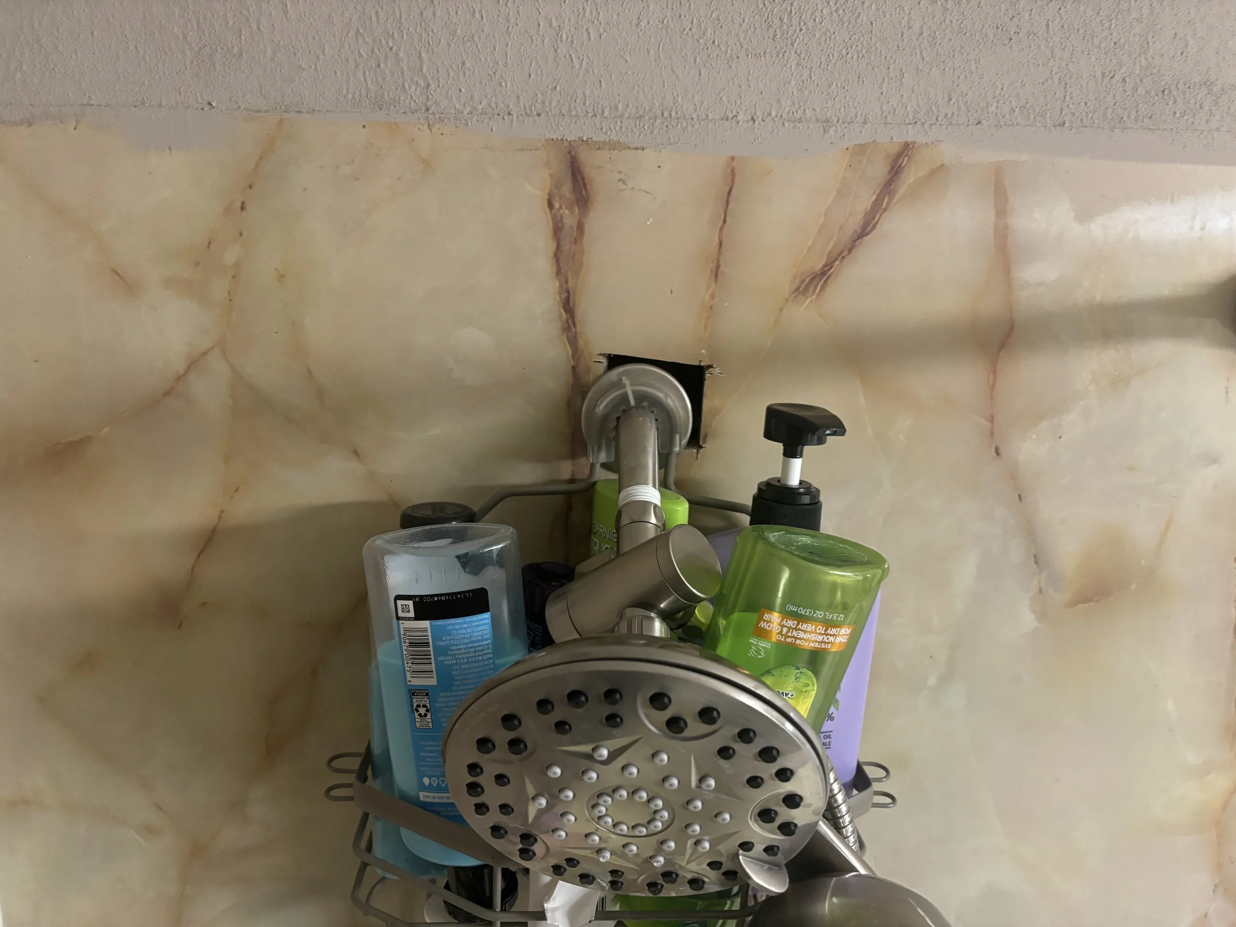 Shower shelf with shampoo, conditioner, body wash, and a showerhead in a bathroom with beige marble walls.