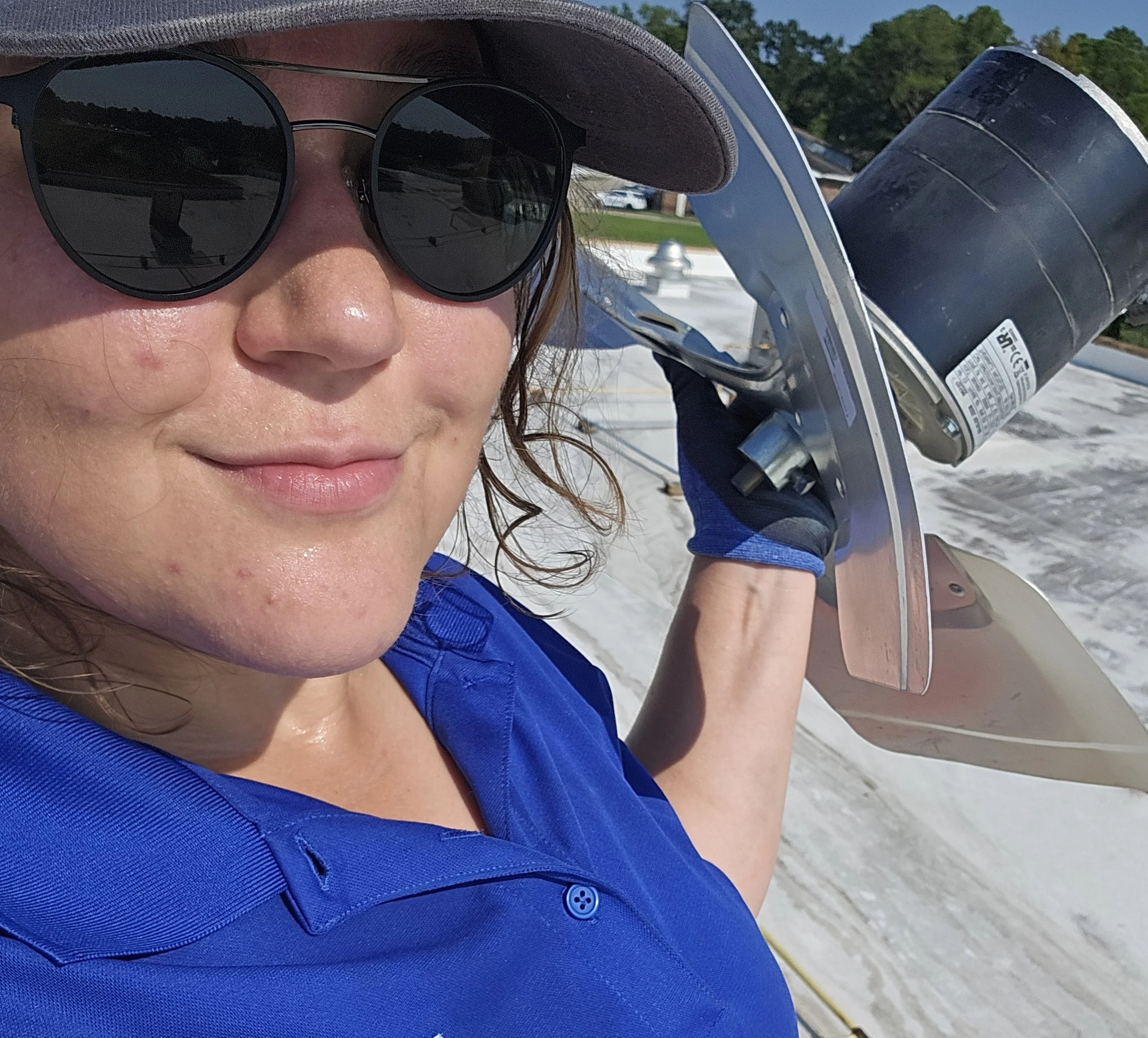A woman wearing sunglasses, a wide-brimmed hat, and a blue shirt, holding a squeegee and a can of spray paint on a rooftop.