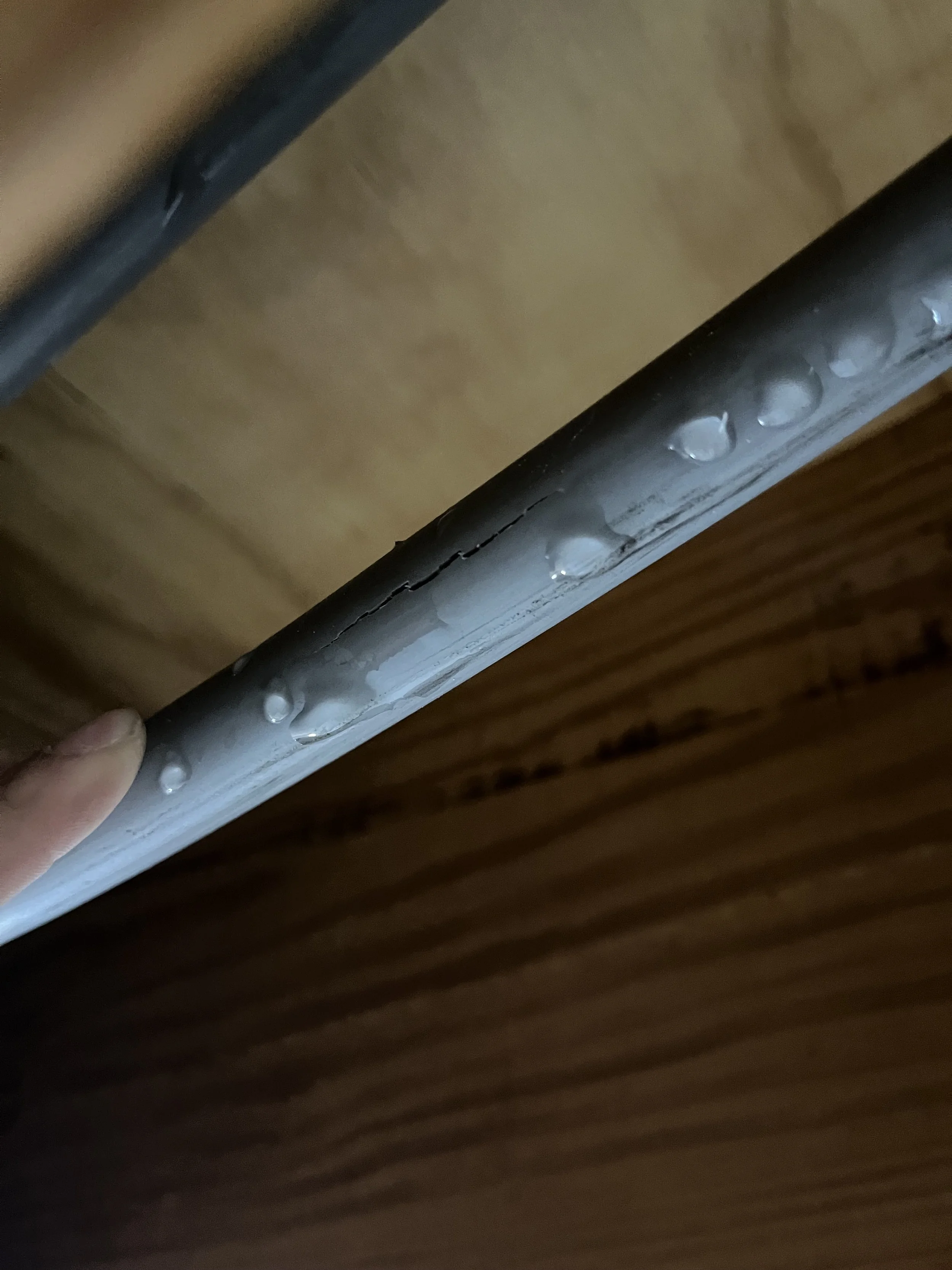 Close-up of a metal pipe with water droplets on its surface, held by a person's hand against wooden and beige background.