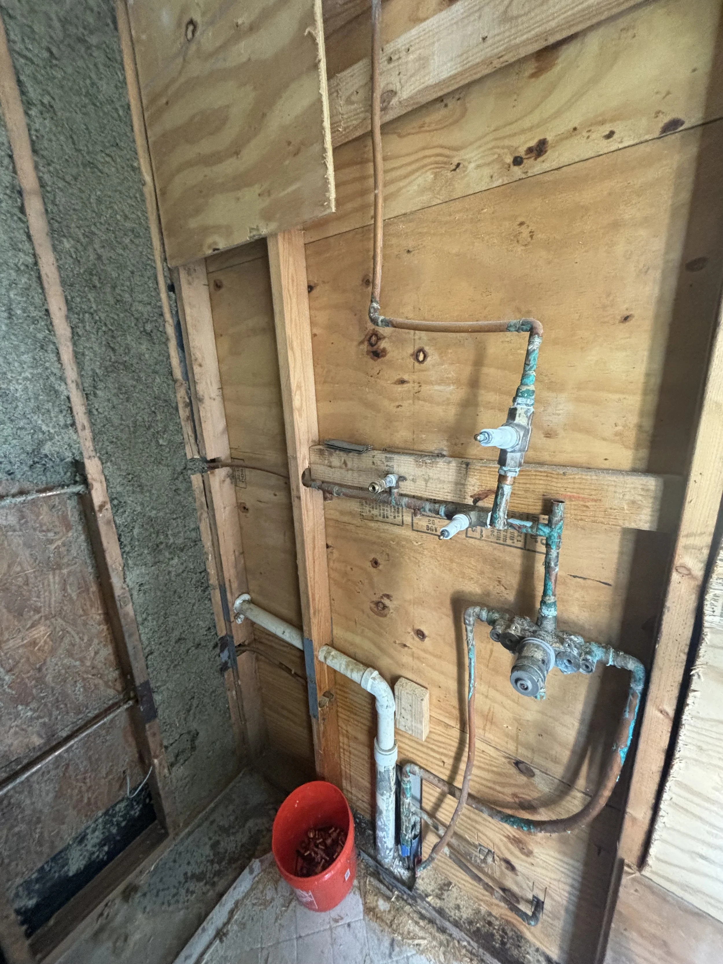 Exposed plumbing pipes on a wooden wall with a bucket of debris on the floor.