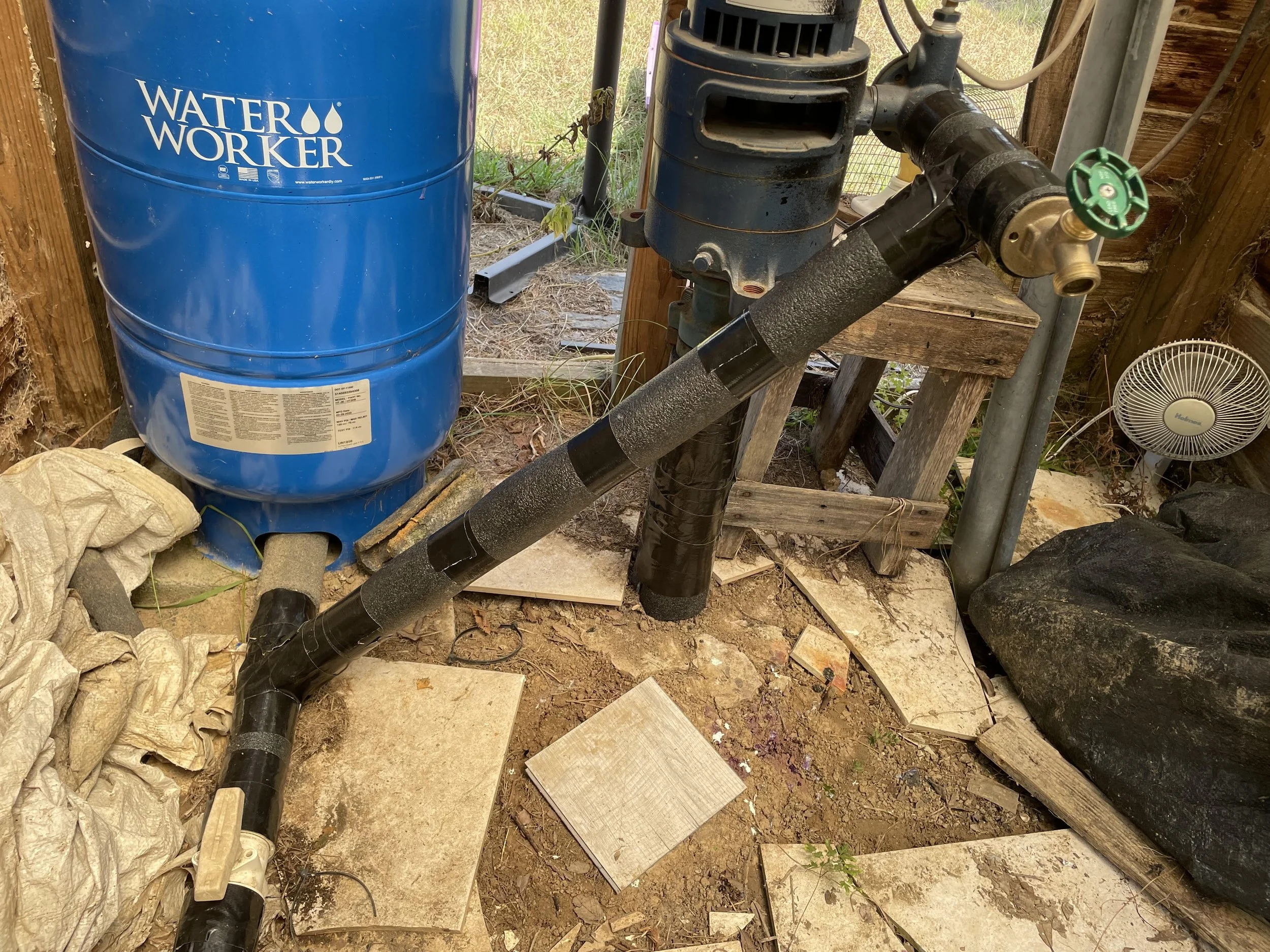 A water filtration system installed next to a wooden fence, featuring a large blue water storage tank labeled 'Water Worker,' connected to black pipes and a pump, with some tools and materials around.