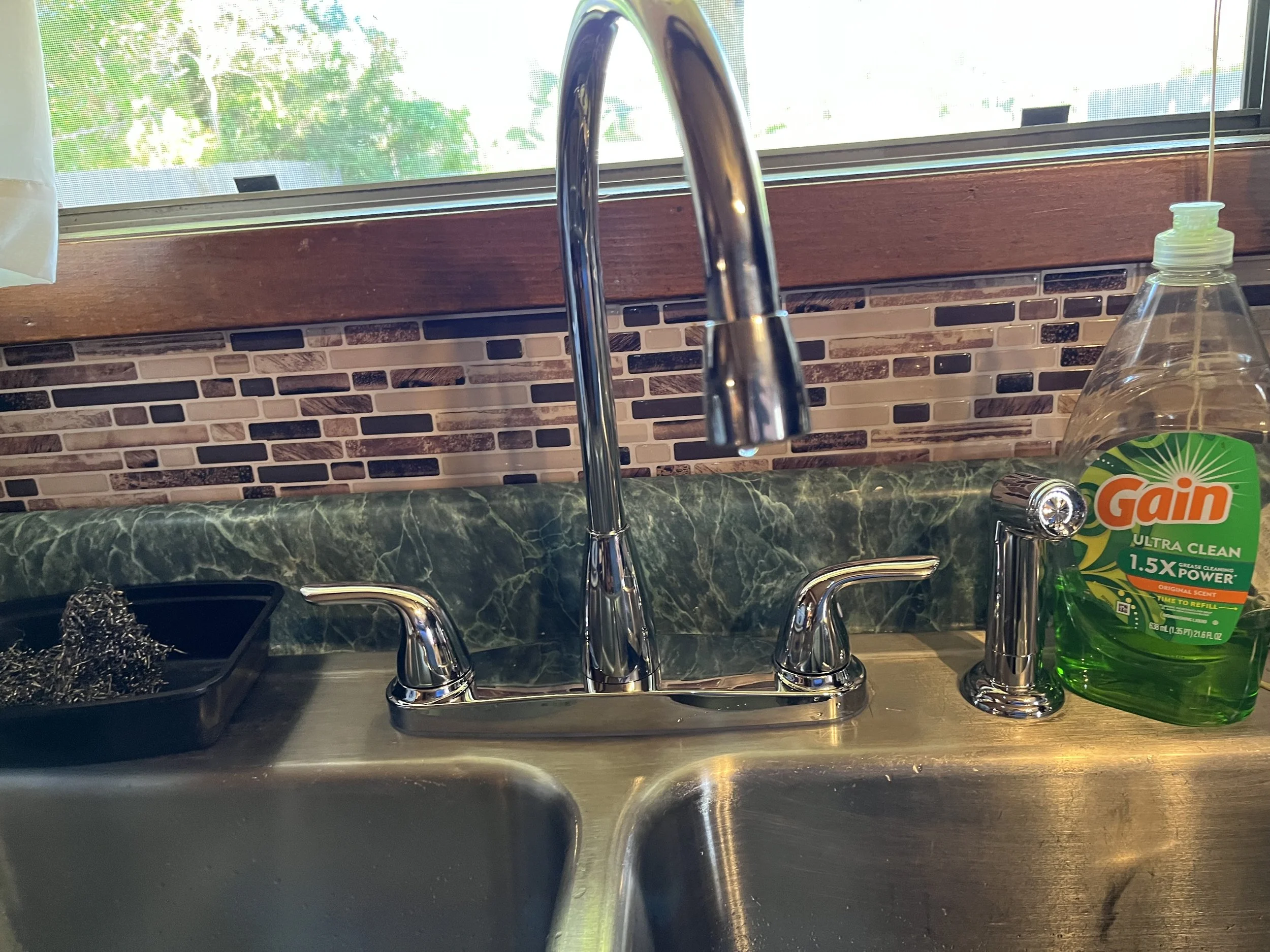 Kitchen sink with two chrome faucets, a green marble countertop, a dish soap bottle labeled Gain, and a black yarn ball holder. A window with a view of trees is in the background.
