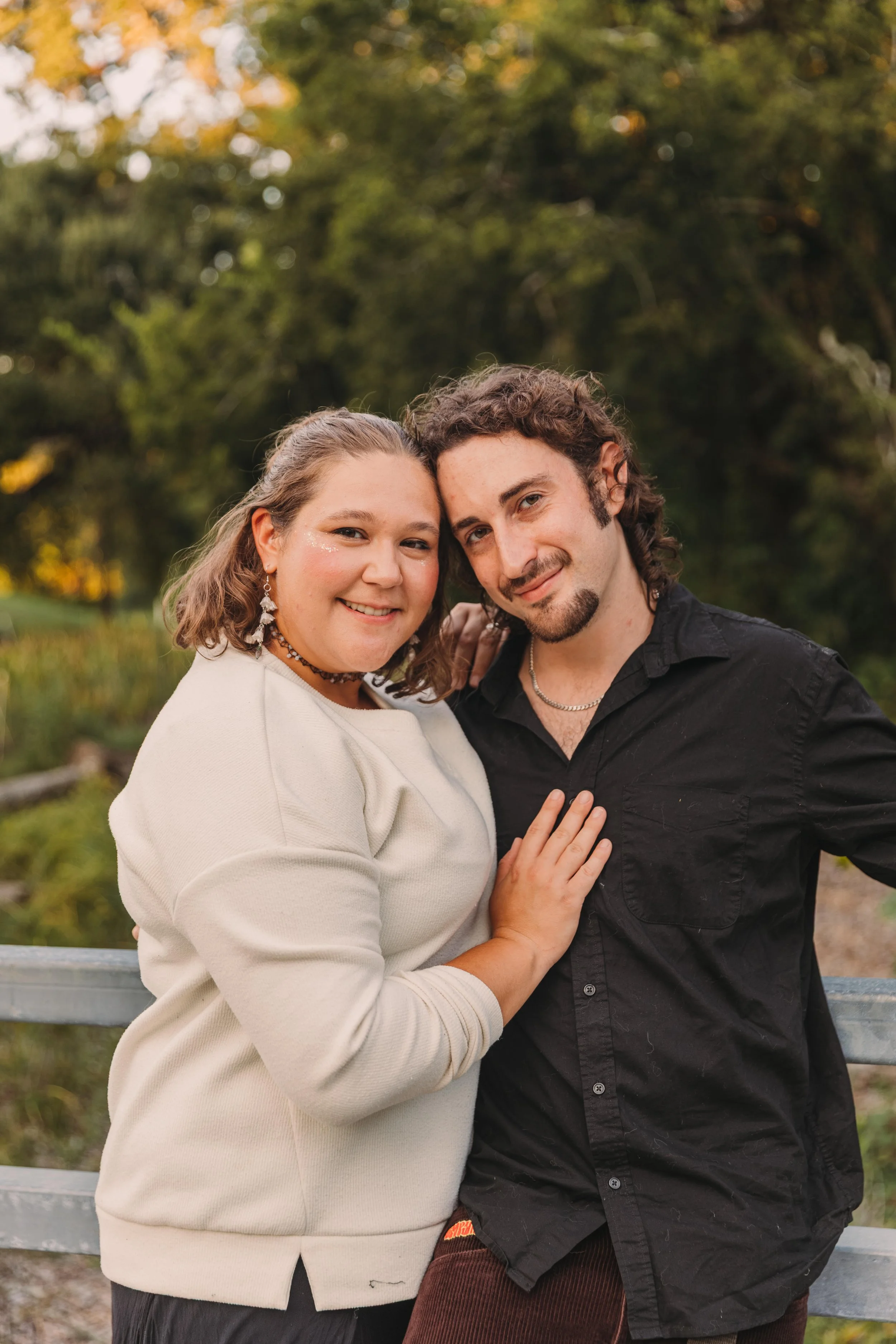 A smiling woman and man standing close together outdoors during autumn, with trees in the background. The woman wears a cream-colored sweater and jewelry, and the man wears a black shirt and necklace.
