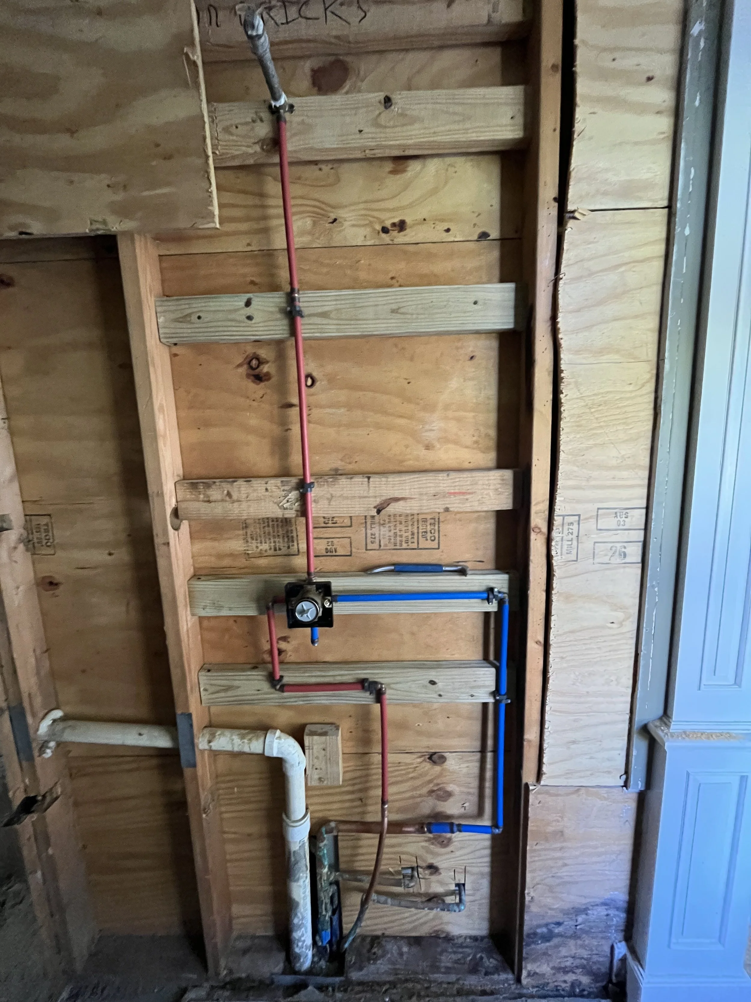 Plumbing pipes, including copper and PVC, installed on a wooden wall frame, with a pipe cutter tool attached to one of the copper pipes.