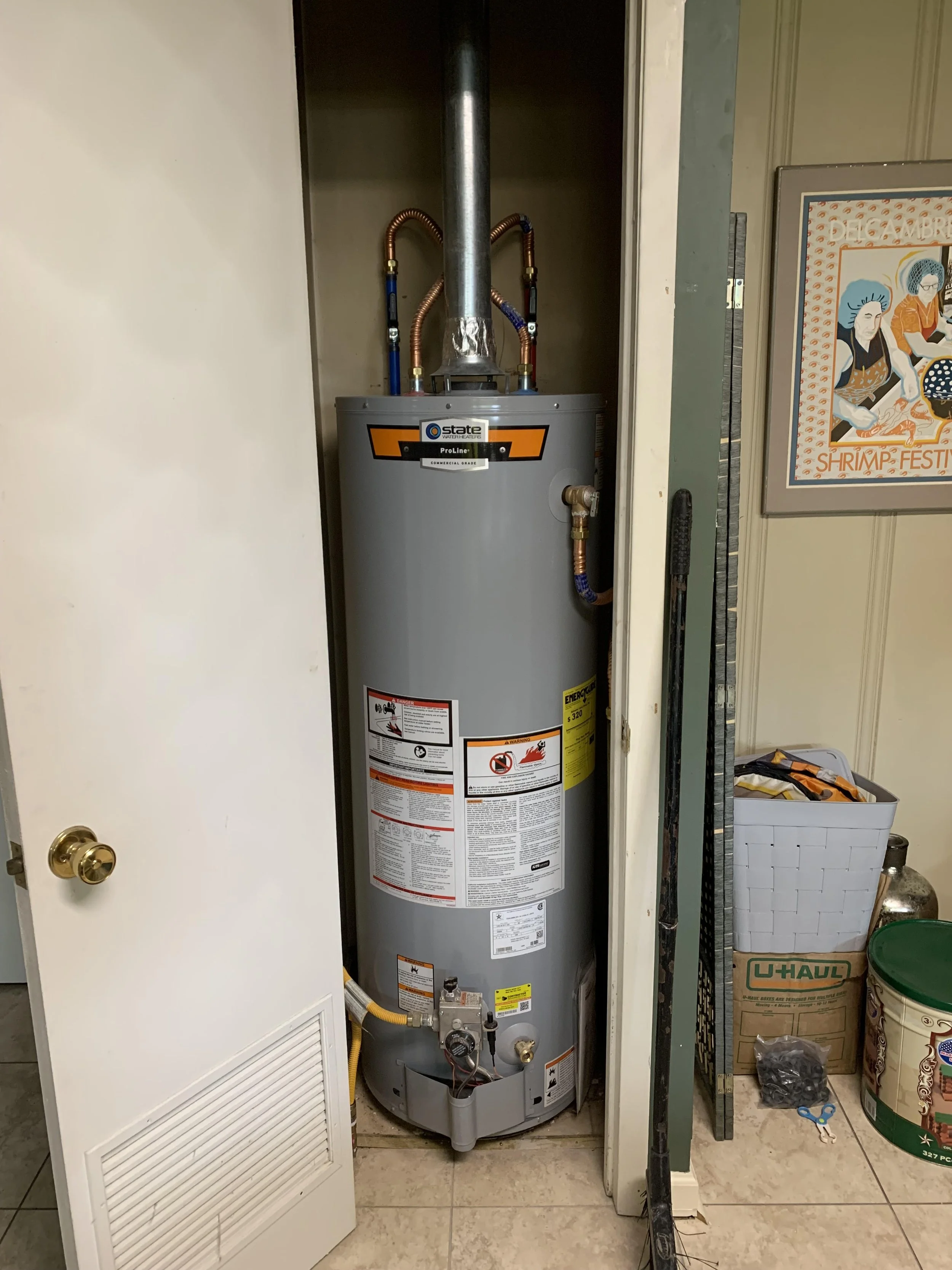 A gray water heater in a small closet next to a partially open door, with pipes connected at the top and labels on the front.