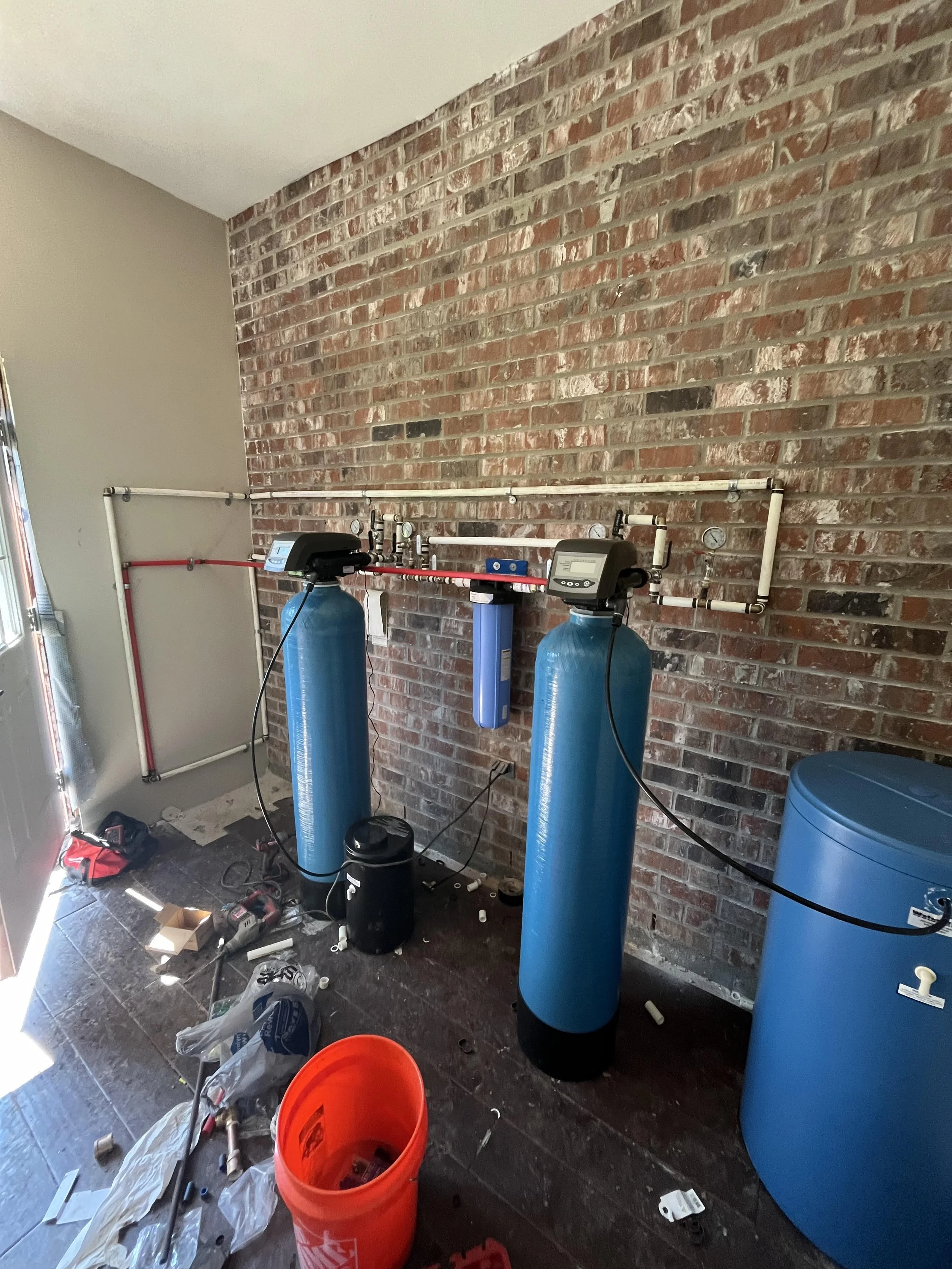 A newly installed water softener system with two large blue tanks and a water filter on a brick wall, with various tools and equipment scattered on the floor.