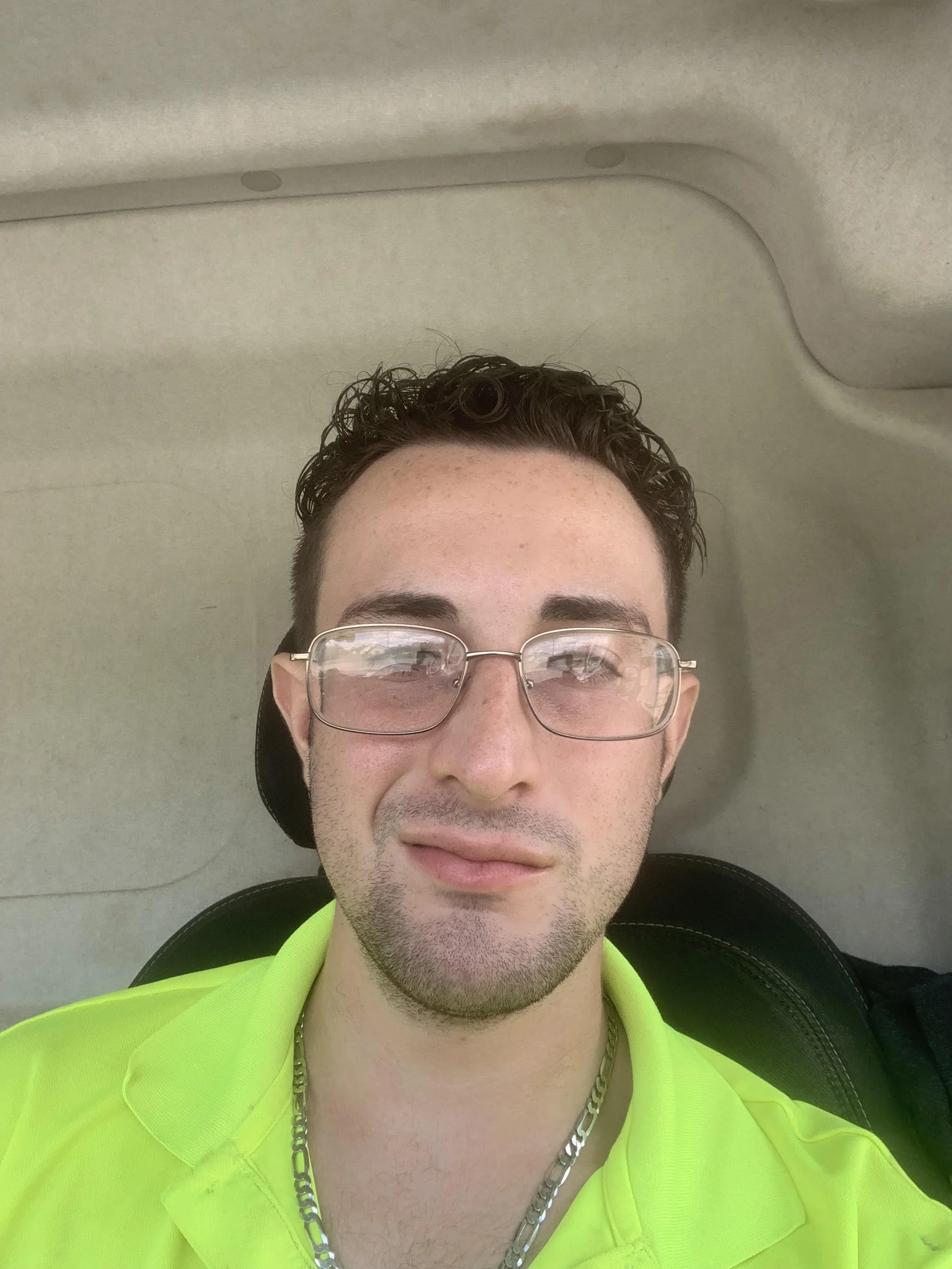 A young man with brown curly hair, glasses, and wearing a neon yellow shirt, taking a selfie inside a vehicle with a beige interior ceiling.