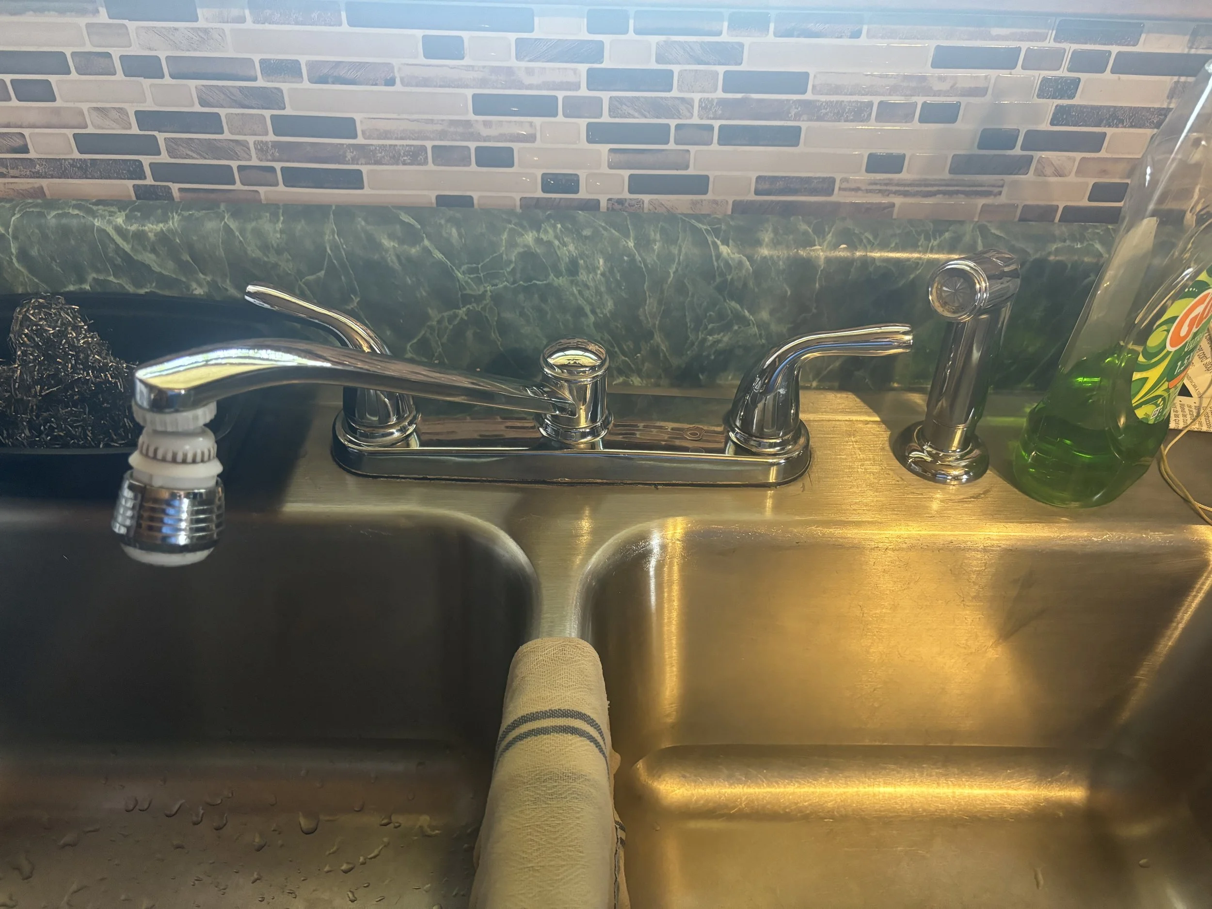 Kitchen sink with a dual-handle faucet, a green soap bottle on the right, a steel scrubber on the left, and a paper towel hanging over the sink in front. The background features a brick-patterned backsplash.