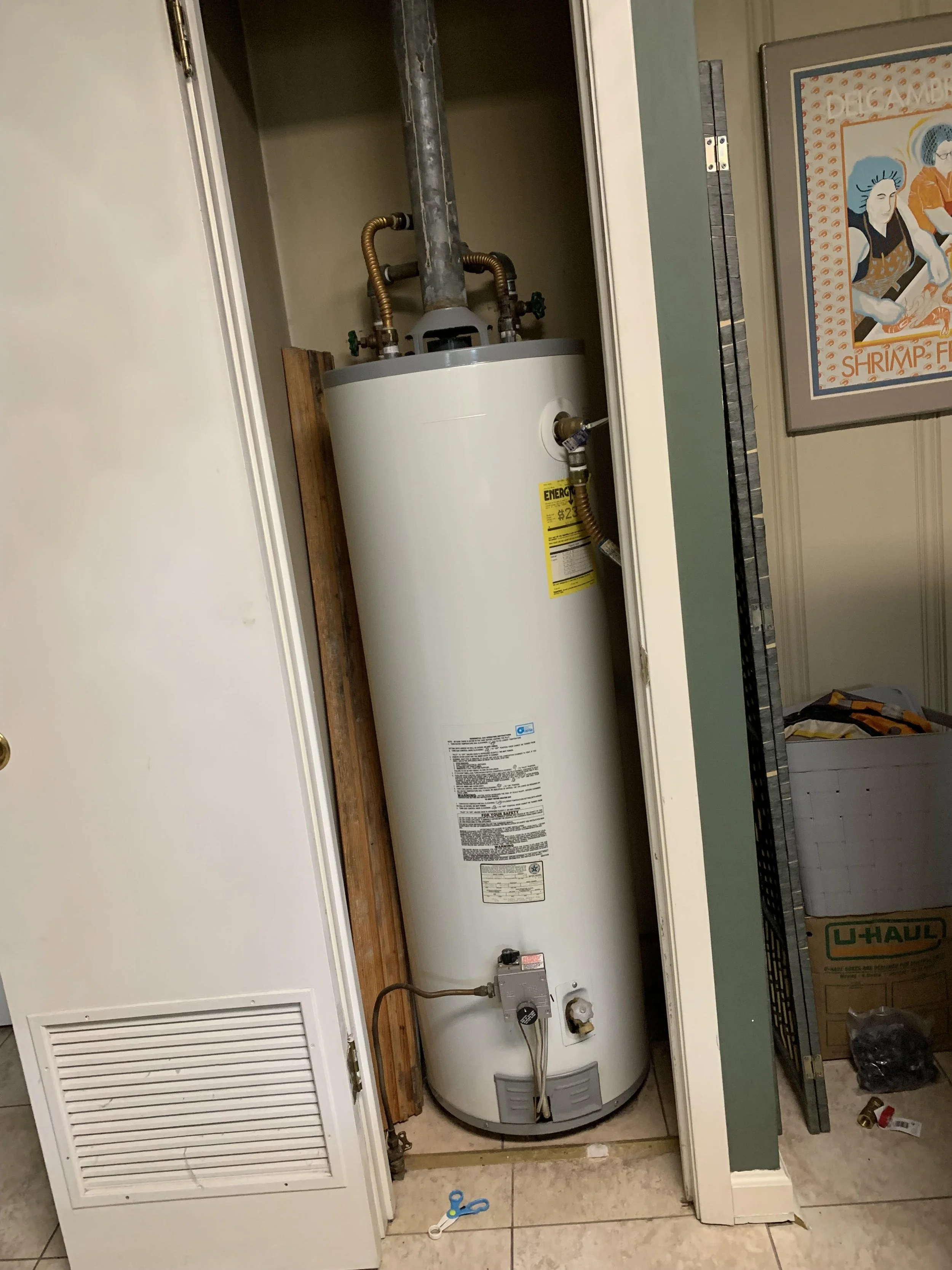 A white water heater installed in a small closet space with copper pipes on top, a yellow energy efficiency label on the side, and a control valve near the bottom. There is a pair of scissors on the floor in front of it.