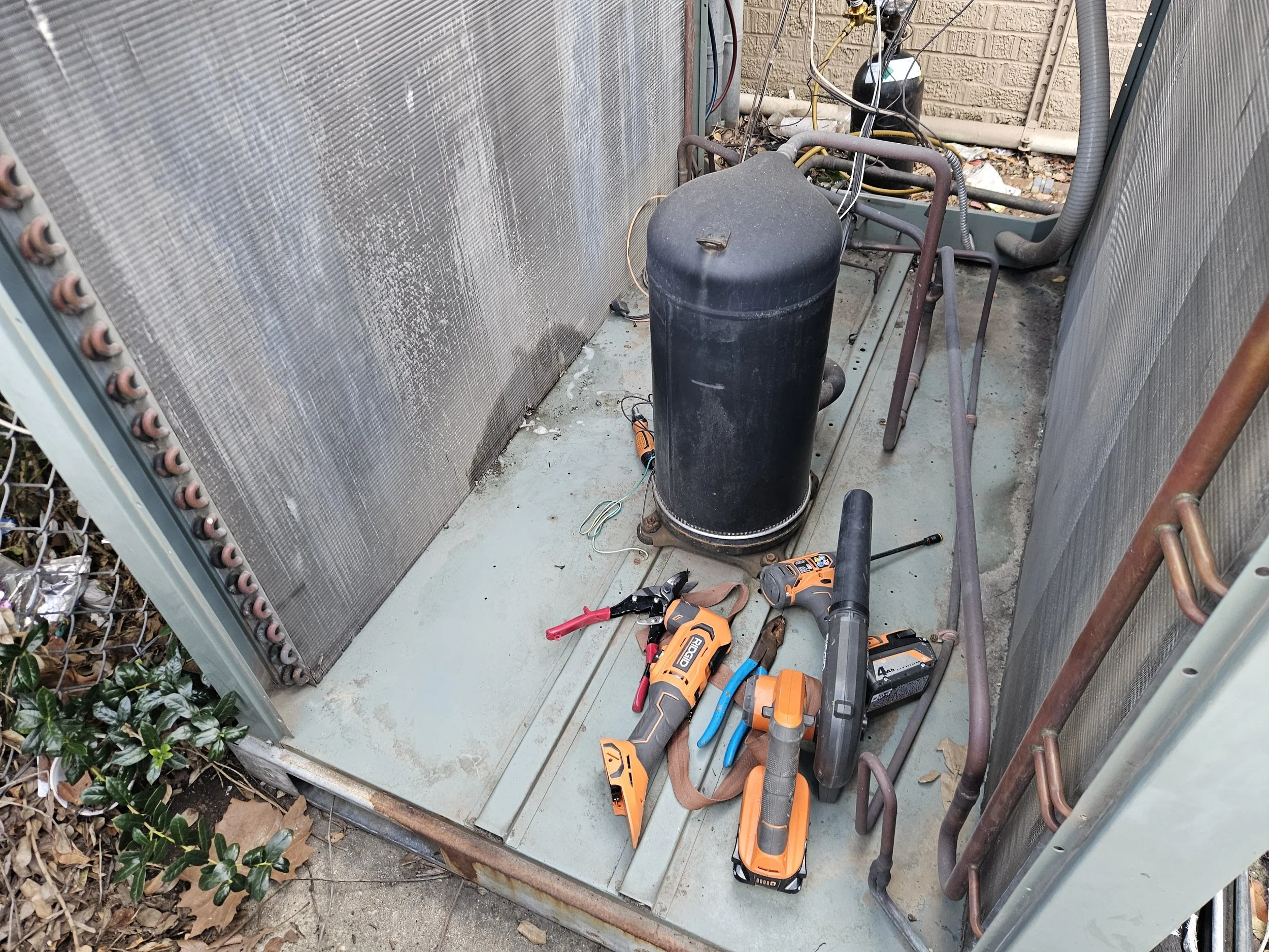 Tools and equipment used for HVAC or refrigeration work, including a black compressor, a cordless drill, a torch, and miscellaneous tools, are placed inside an outdoor HVAC unit enclosure with exposed wiring and pipes.