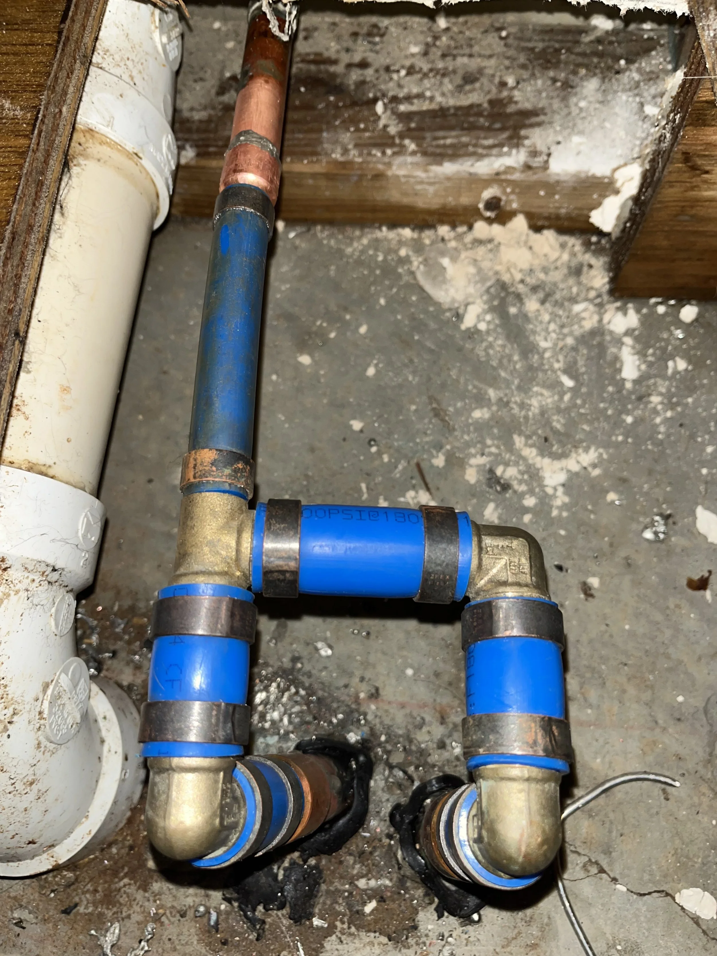 Close-up of new blue and brass plumbing pipes with copper pipes, fittings, and threaded connections on a concrete floor next to a white PVC pipe.