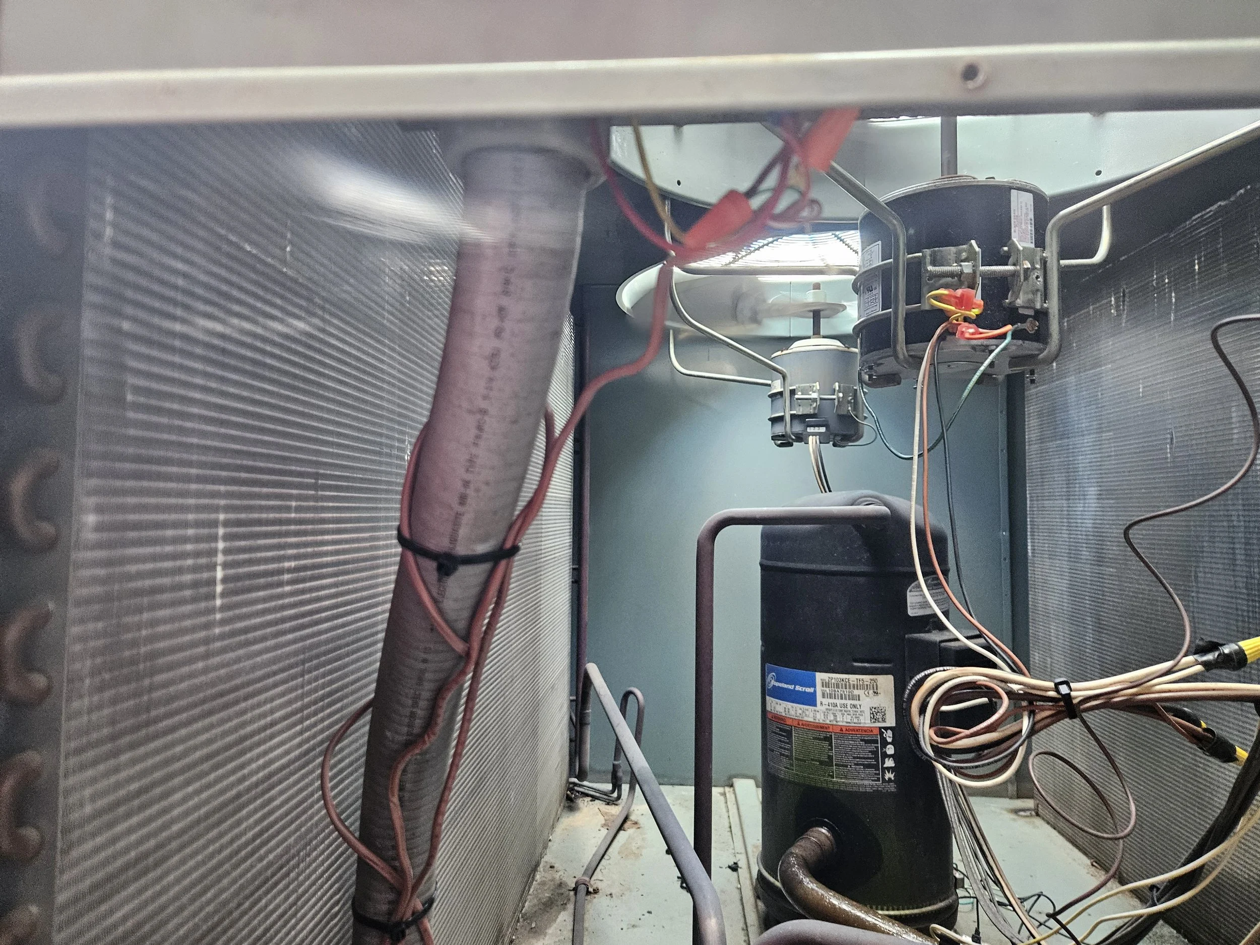 Interior view of an air conditioning unit revealing compressor, electrical wires, pipes, and metal fins.