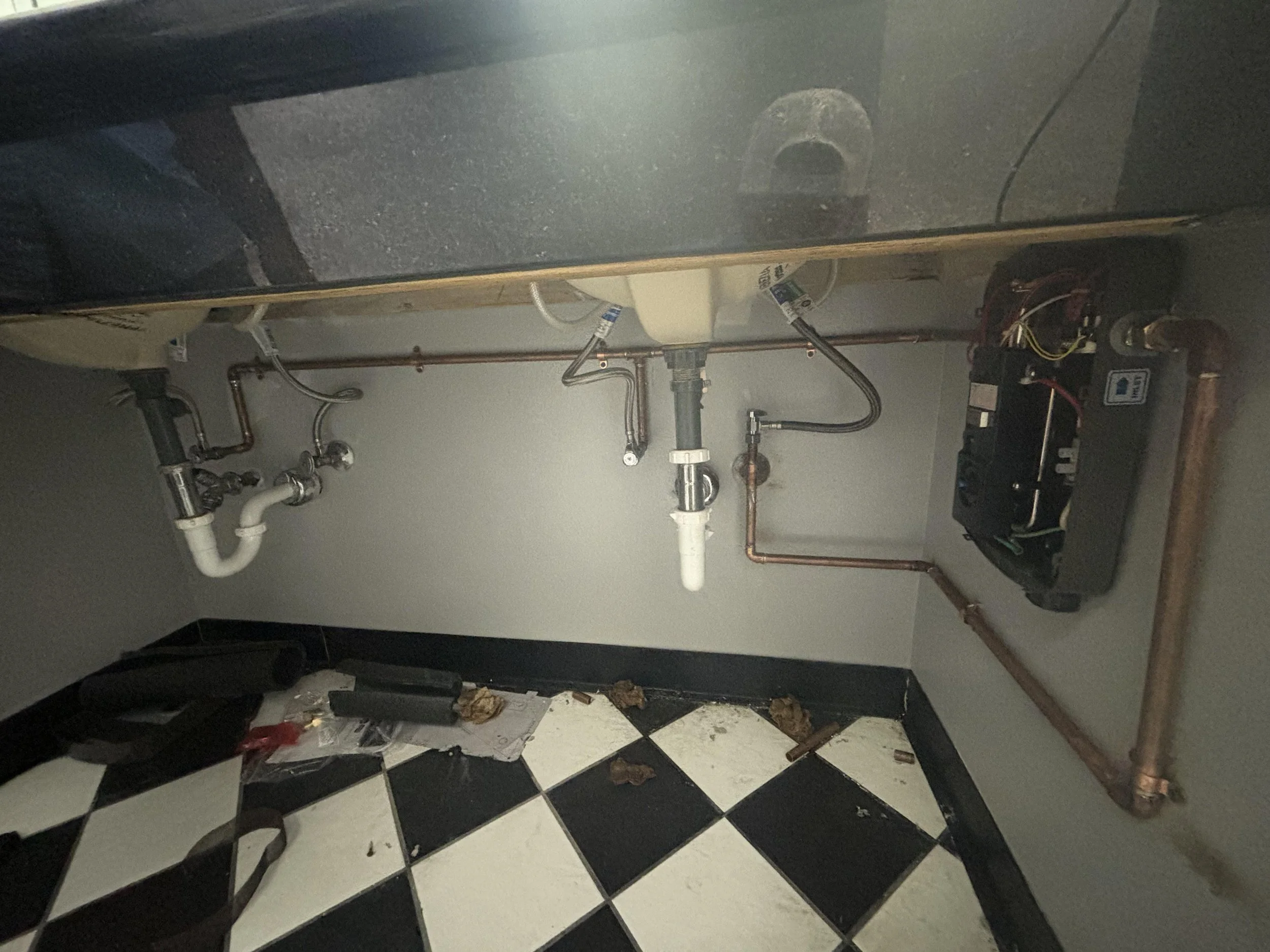 Under-sink plumbing with pipes and a water filter, black and white checkered floor, cleaning supplies and debris on the floor.