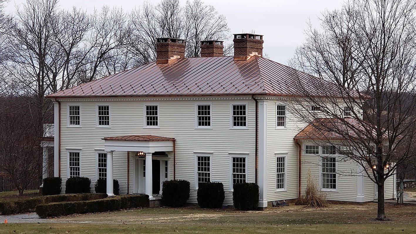 White two-story house with a copper roof, multiple windows, and a small porch supported by two white columns, surrounded by leafless trees and shrubs.