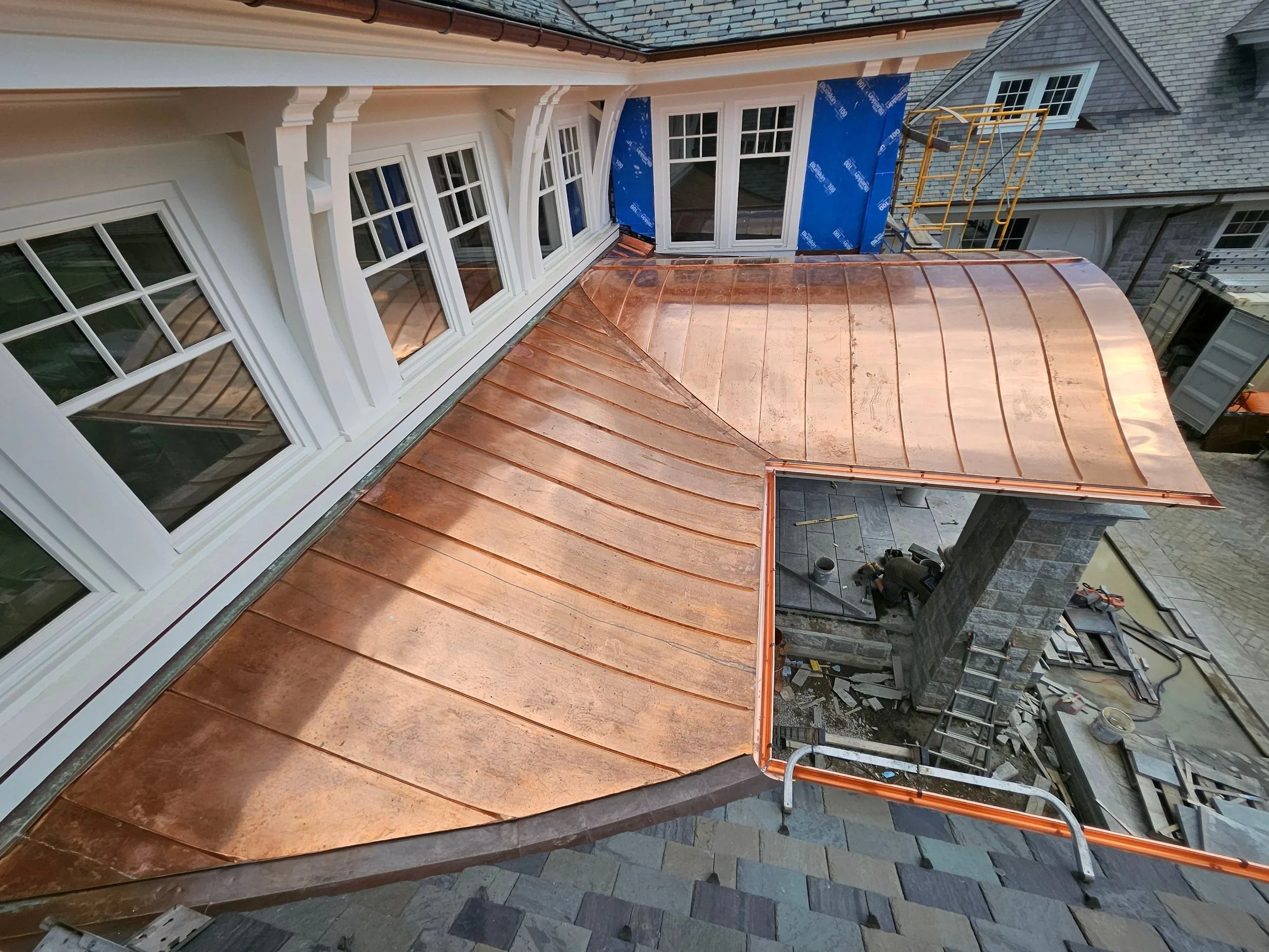 View of a residential roof under construction, showing copper roofing material, white window frames, and part of the house facade with a brick exterior. Construction scaffolding and tools are visible.