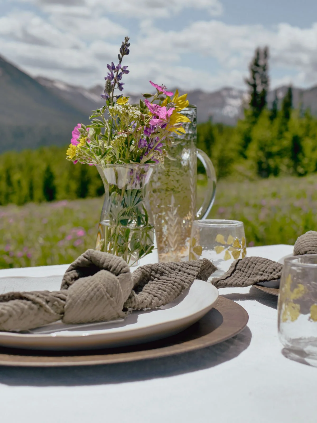 Table set outdoors with plates, glasses, and a vase of wildflowers, with mountains and trees in the background.