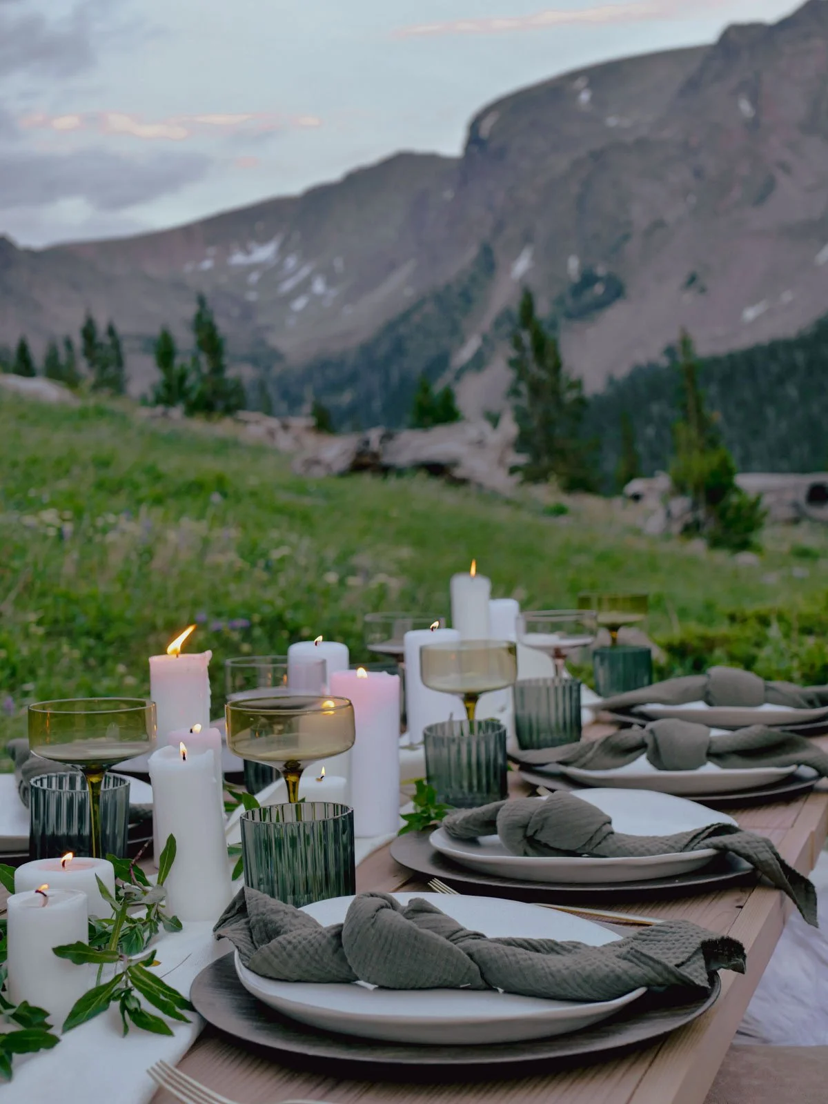 A rustic outdoor dining table set with white and gray tableware, gray cloth napkins, and tall white candles in gold and glass holders, with green foliage accents, against a mountainous landscape background during dusk.
