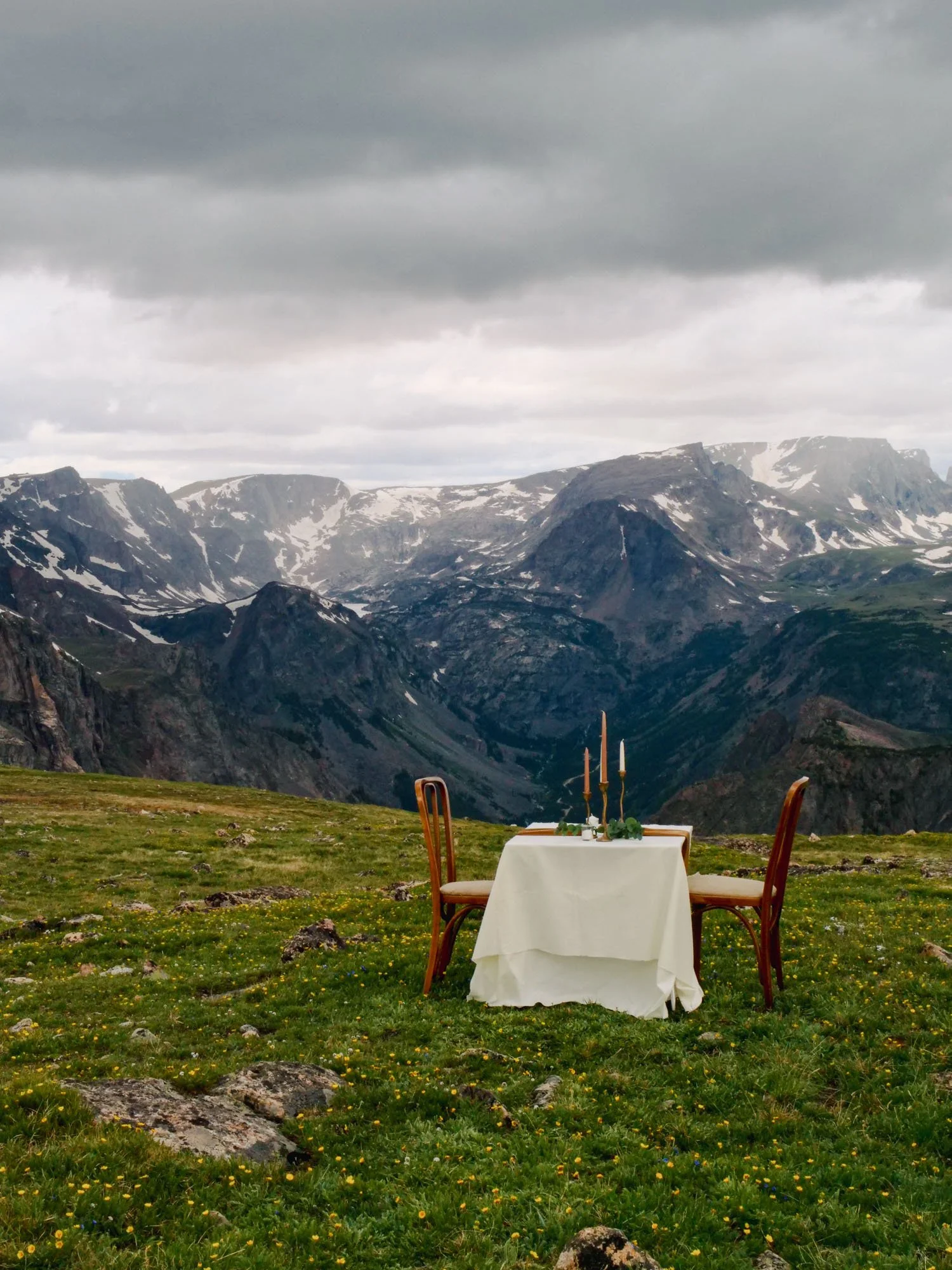 A table set for two with candles, placed outdoors on grassy terrain, with mountain range and cloudy sky in the background.