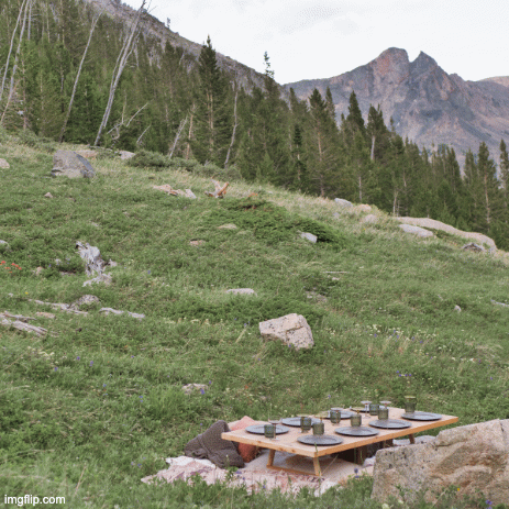 A picnic setup on a wooden table in a grassy mountainous area with trees and mountain peaks in the background.