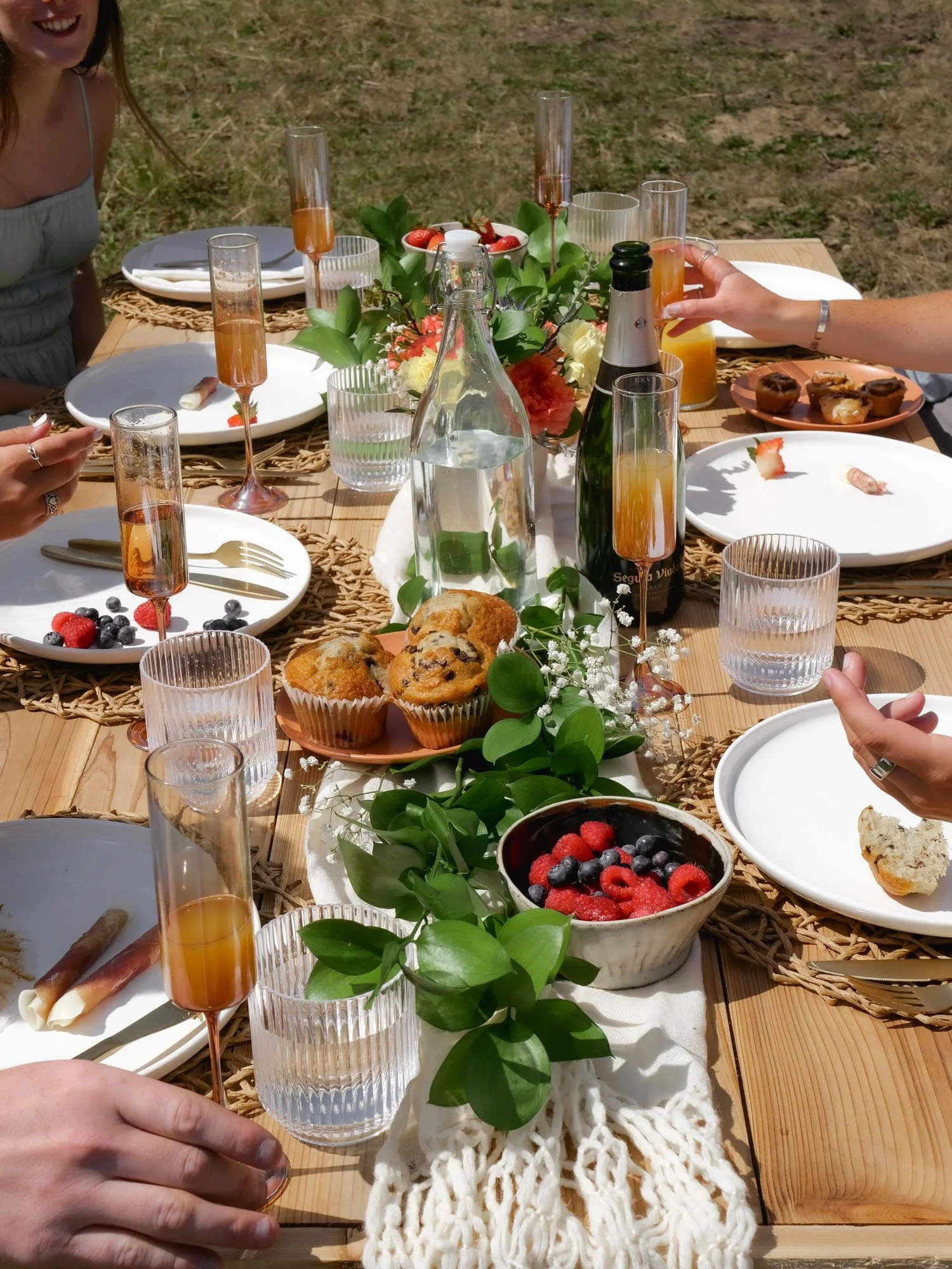 A table set for a gathering with plates, glasses, and food including muffins, berries, and cupcakes, decorated with greenery and flowers, outdoors in sunlight.