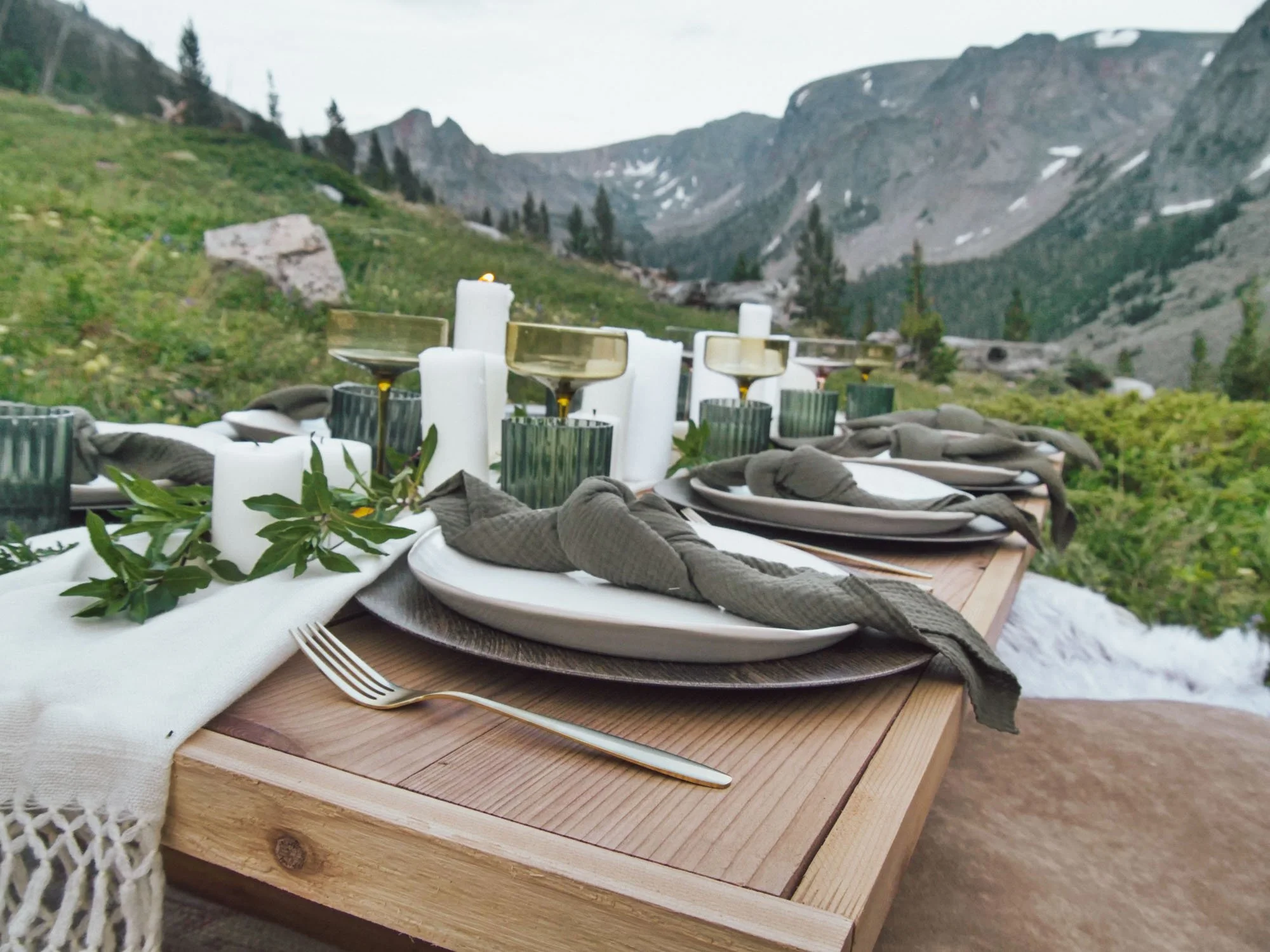 Outdoor mountain dining table set with white plates, cloth napkins, greenery, candles, and glassware, overlooking a mountainous landscape.