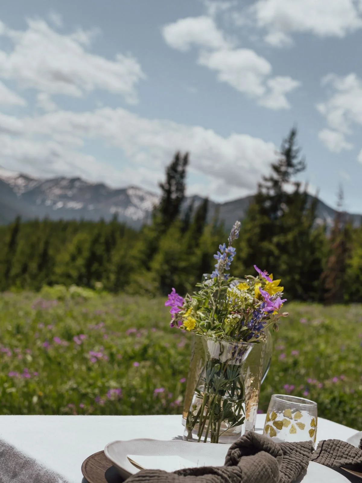 Vase of mixed wildflowers on a table outdoors with mountains, trees, and clouds in the background.