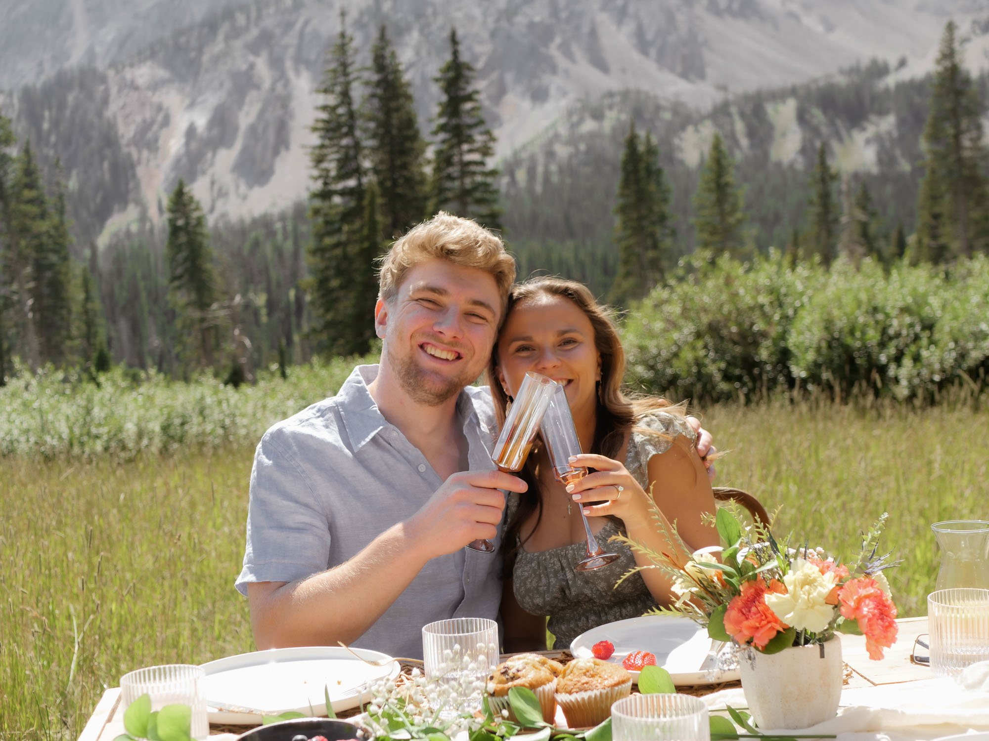 Couple smiling "cheers"ing their champagne flutes. Flowers on table in foreground, tall pine trees and mountains in background.