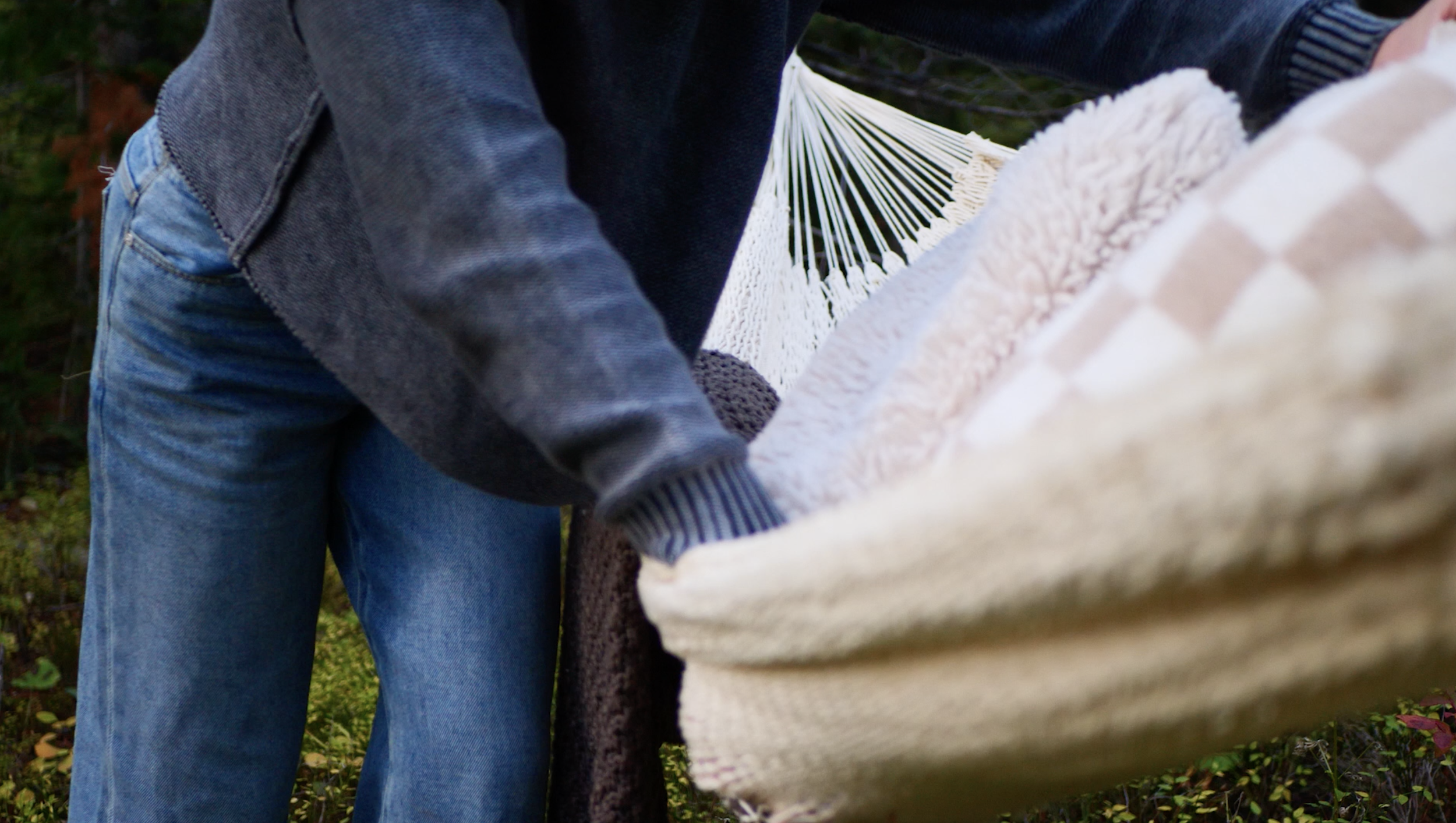 Person in a dark sweater and blue jeans arranging or adjusting a hammock outdoors amid greenery.