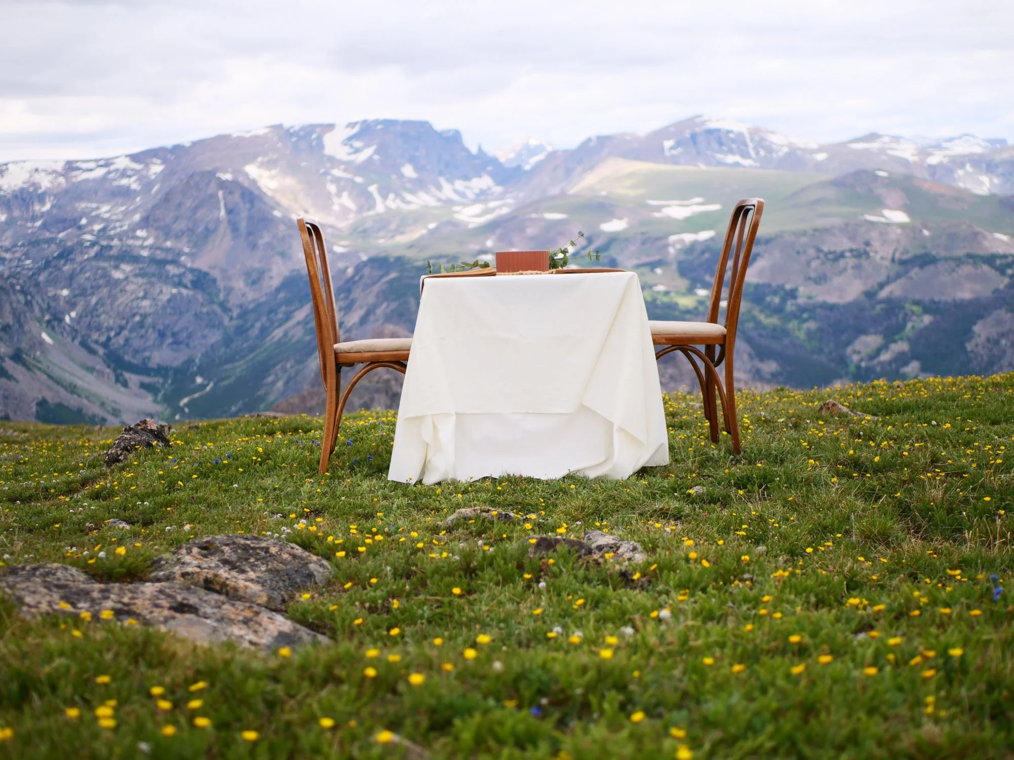 A round table with a white tablecloth and two wooden chairs outdoors on a grassy area with small yellow flowers, mountain landscape in the background.
