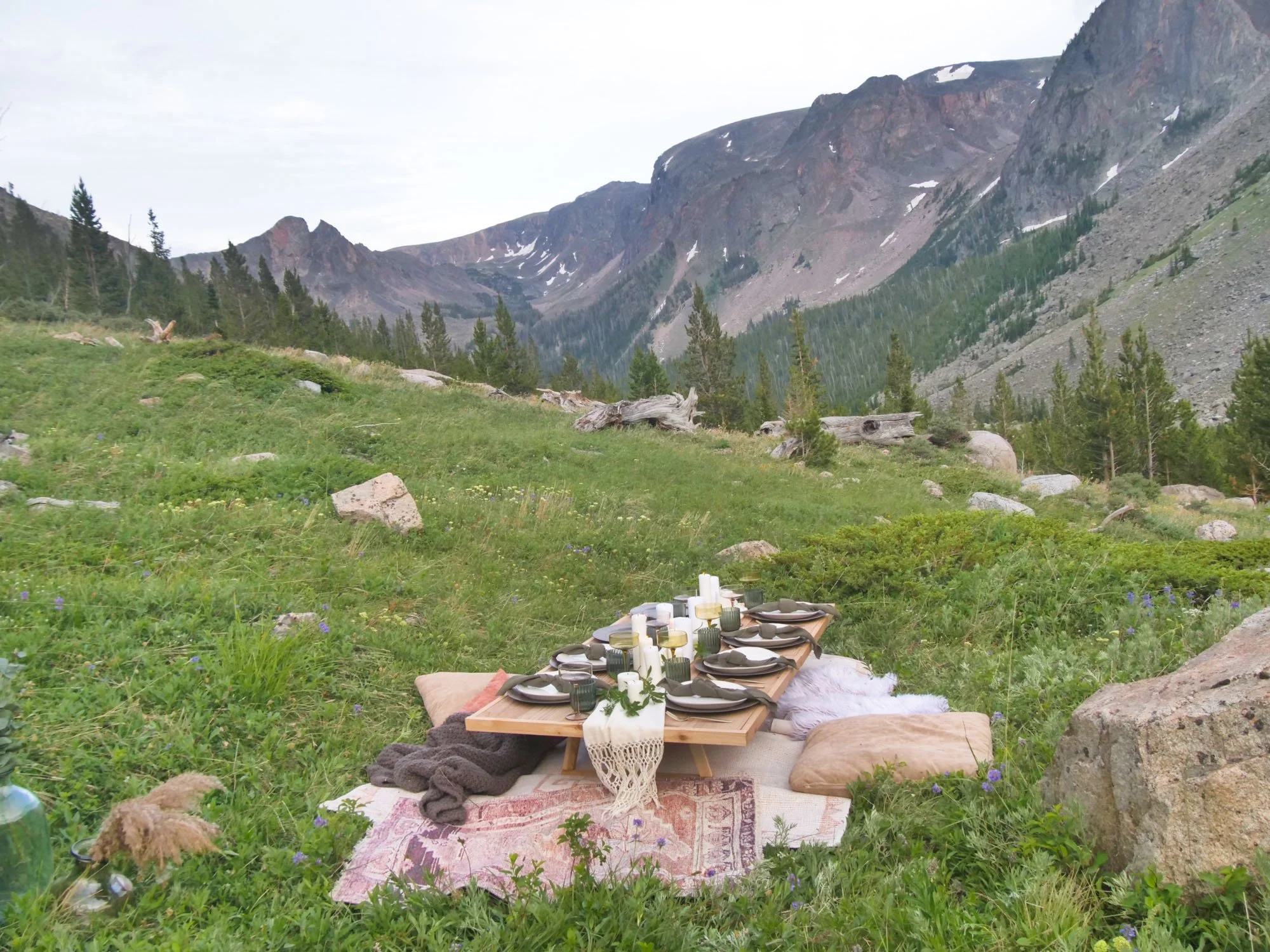 Low tabletop set with candles, pillows, and blankets for a picnic style dinner with plates set for eight people. Table is set in a grassy area with large mountains in the background.